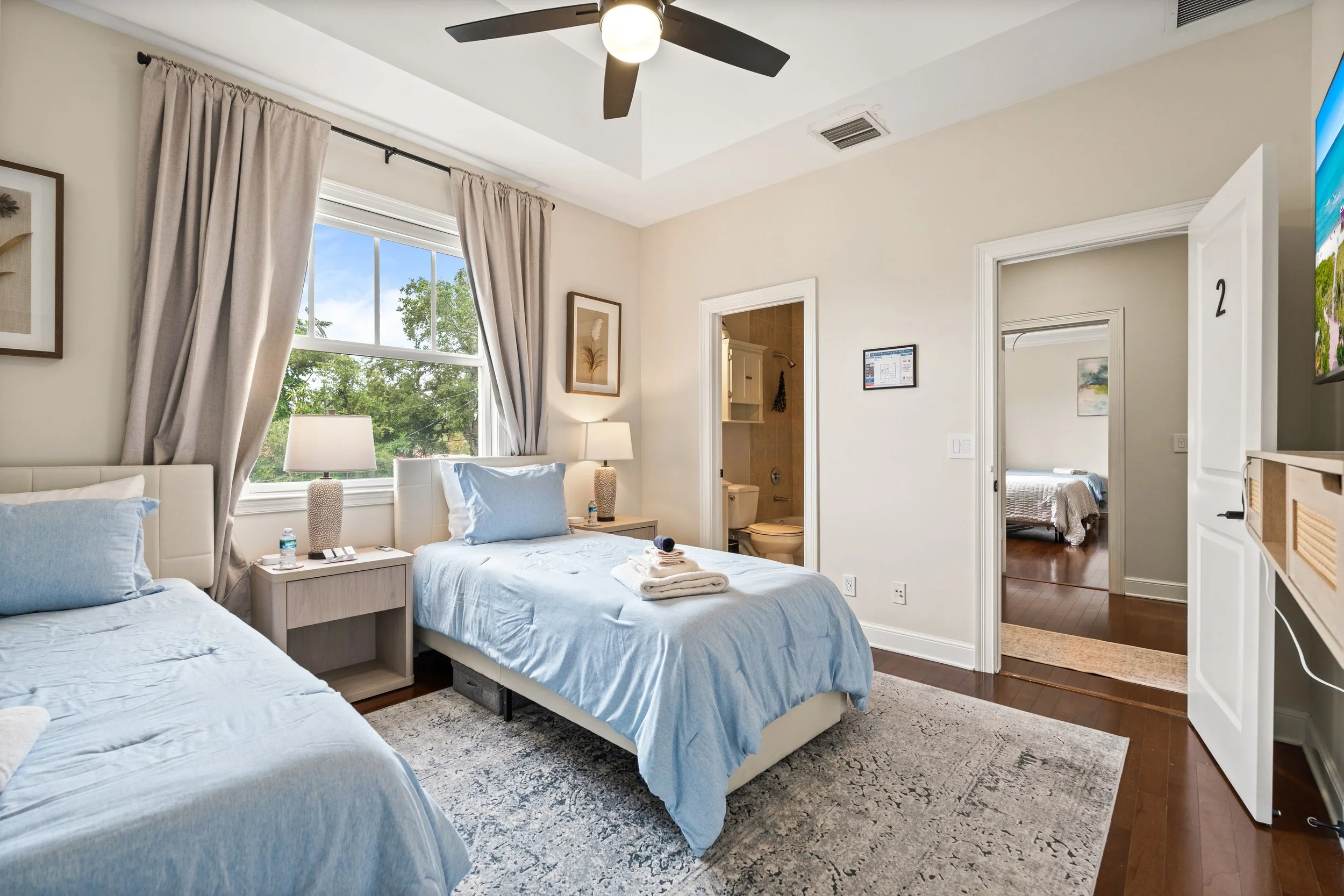 A bedroom with two beds with light blue bedding, a nightstand with two lamps, and a partially open door leading to a bathroom and another bedroom.