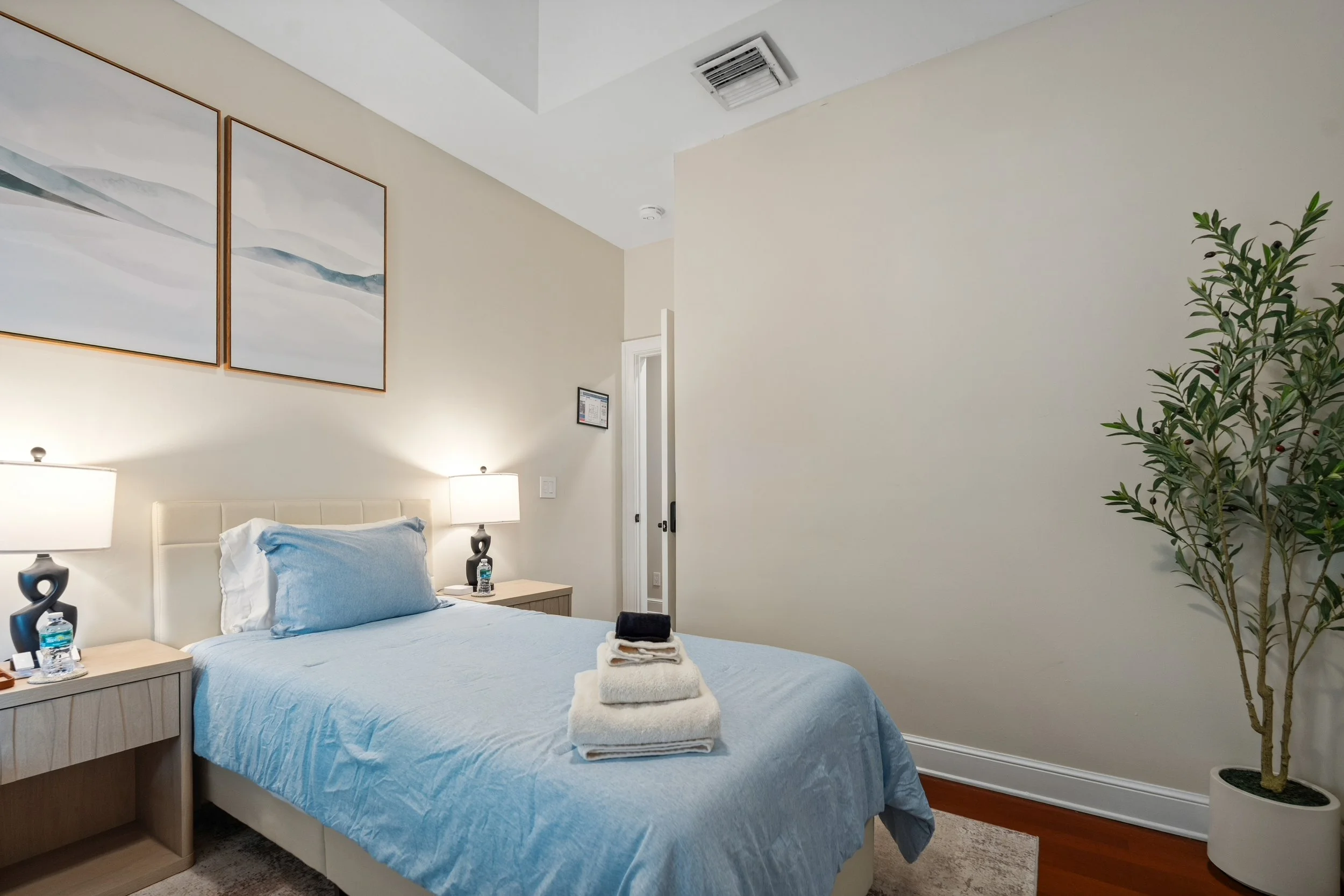 A cozy bedroom with a bed covered in a light blue comforter and pillow, two nightstands with lamps, and neatly folded towels on the bed. There is a tall green potted plant to the right and framed artwork on the wall above the bed. The room has off-wh