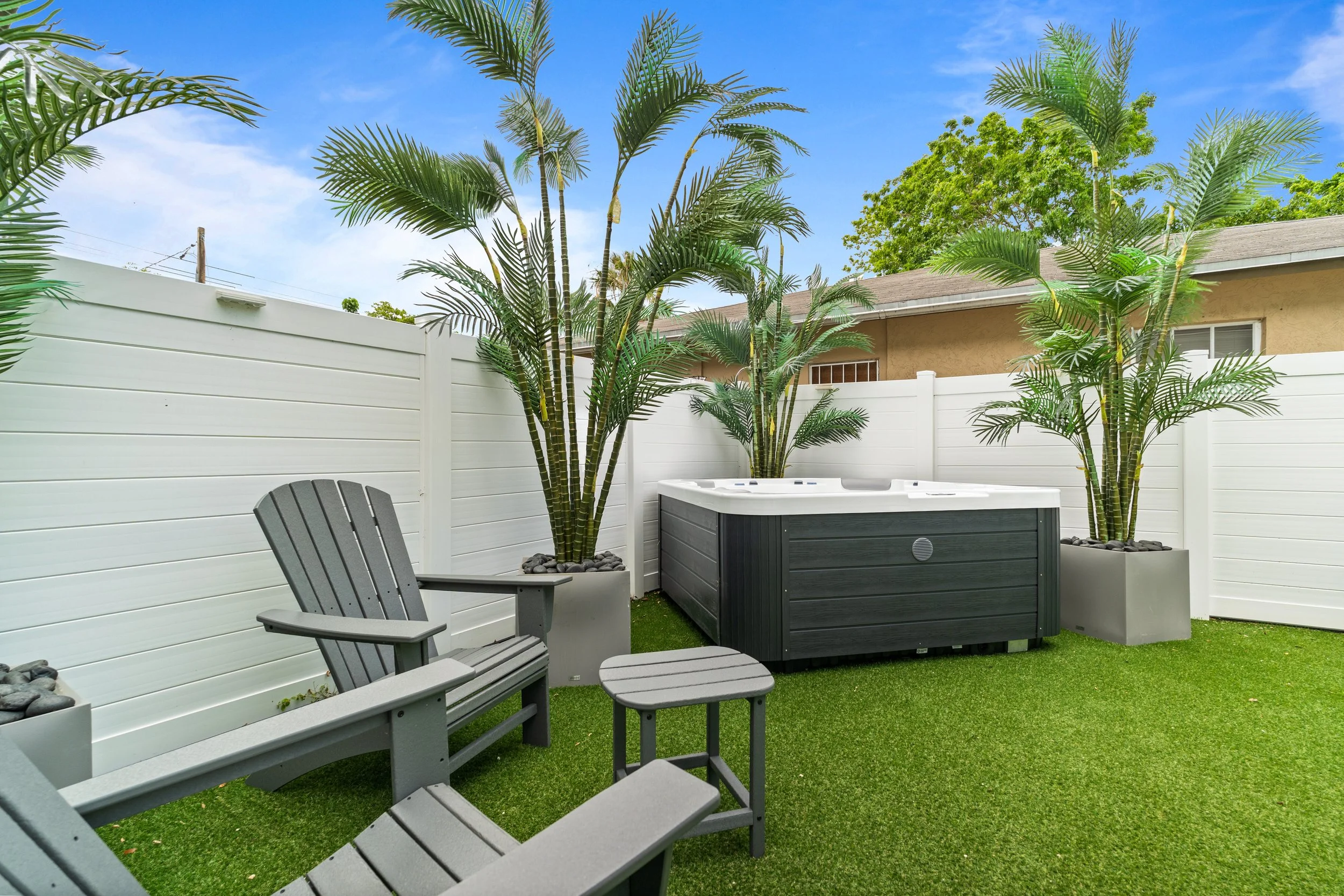A backyard with a white privacy fence, two potted palm trees, gray Adirondack chairs, and a hot tub on artificial grass, with a yellow house and trees in the background under a blue sky.