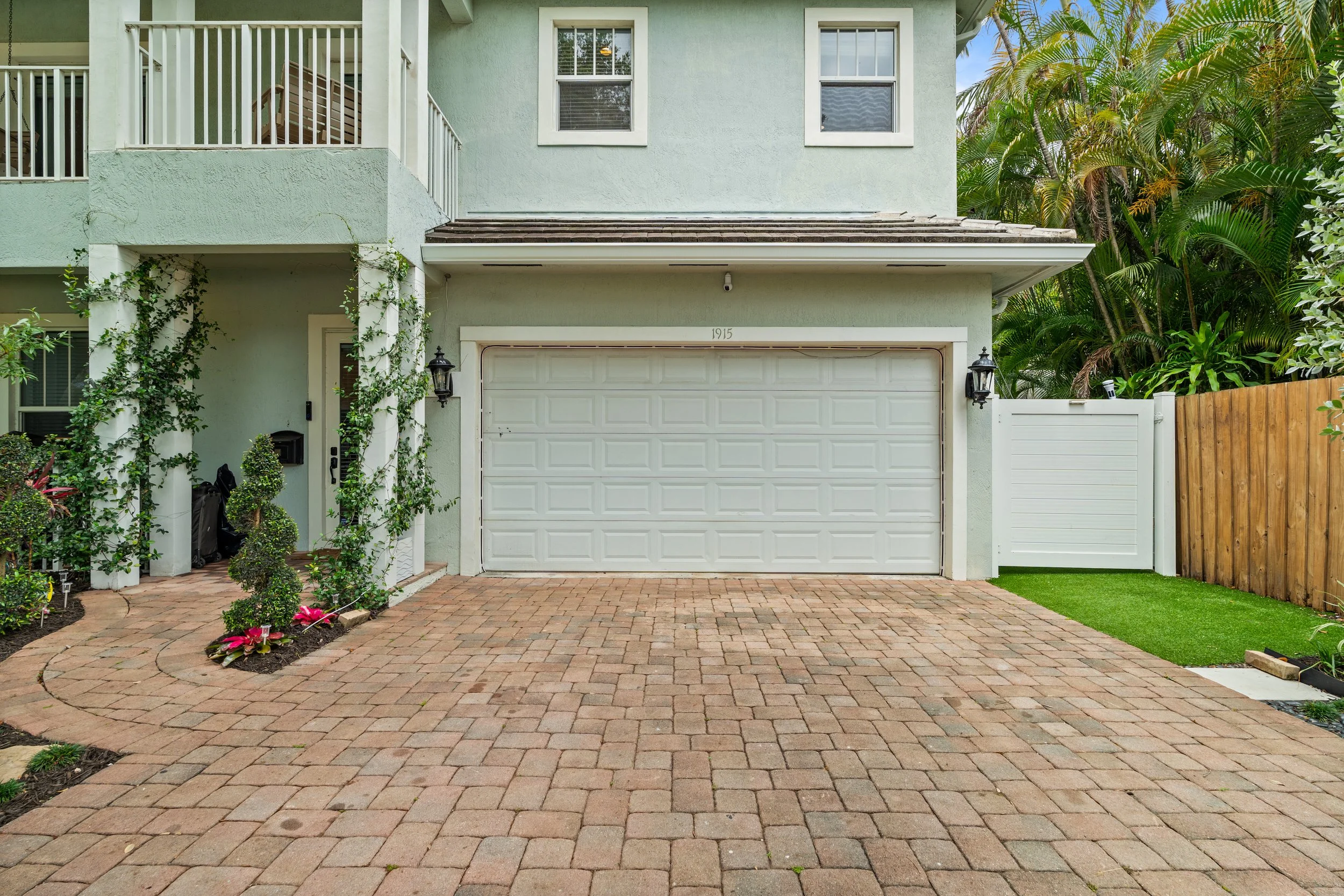 Front view of a two-story house with a white garage door, light green exterior walls, and decorative plants and flowers in the garden. Green trees are visible on the right side of the house.