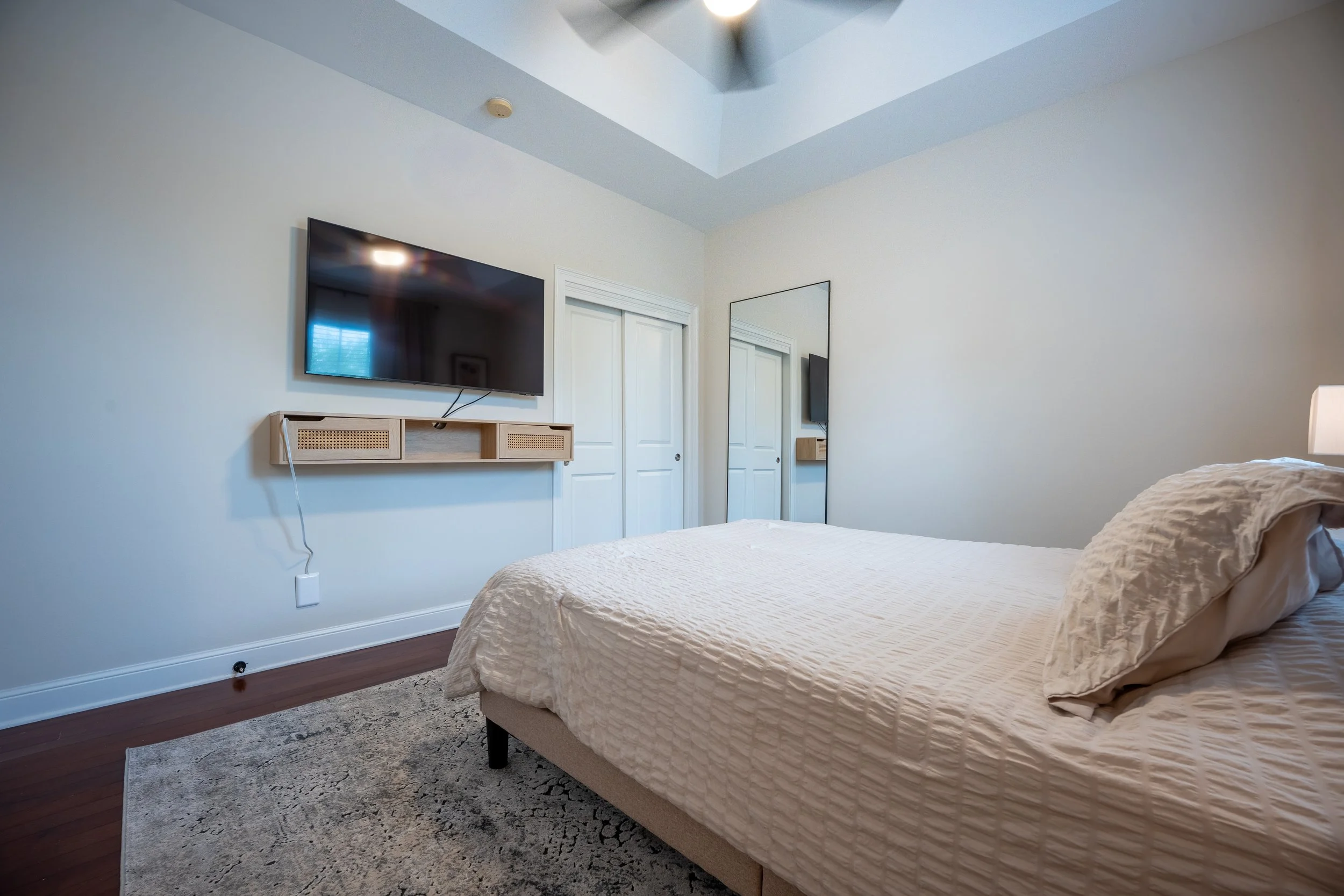 A bedroom with a bed, two closed closet doors, a wall-mounted flat-screen TV, a long narrow mirror, and a ceiling fan with a light.
