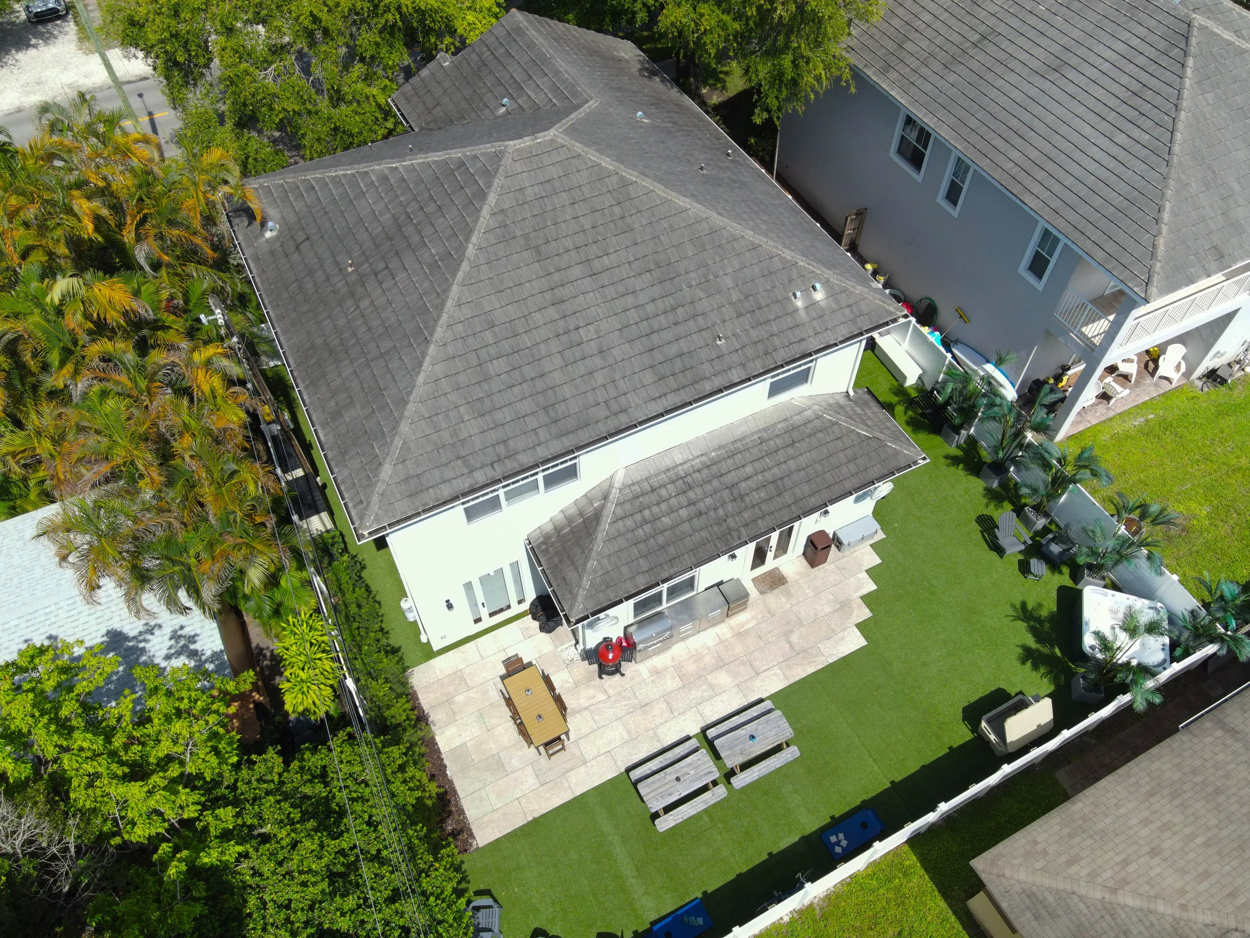 An aerial view of a house with a gray roof, surrounded by trees, a backyard patio with outdoor furniture, and a lawn with some benches and a barbecue grill.