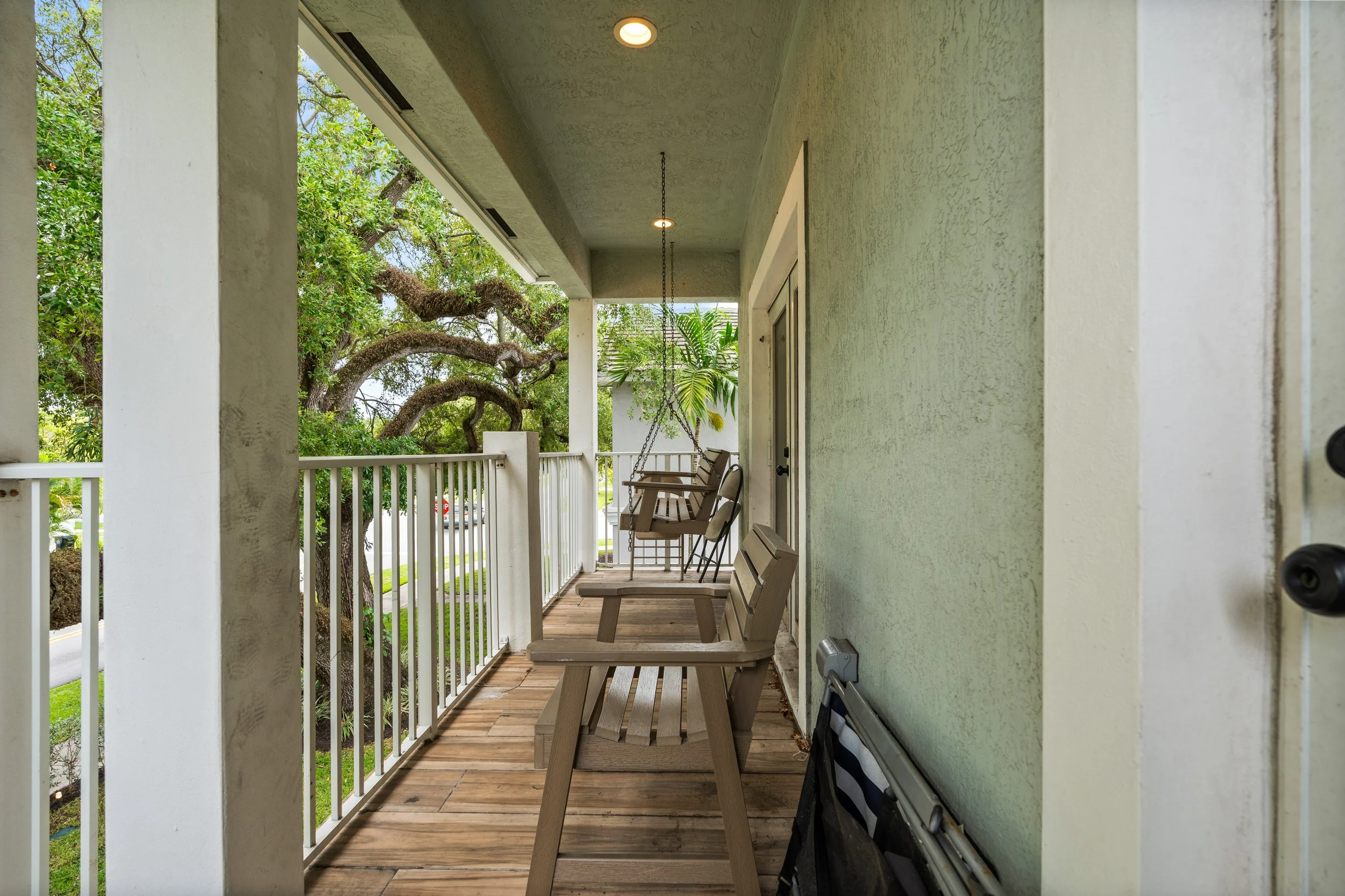 Covered porch with wooden floor, green stucco wall, beige wooden benches, hanging porch swing, and white railing, overlooking green trees and neighborhood street.