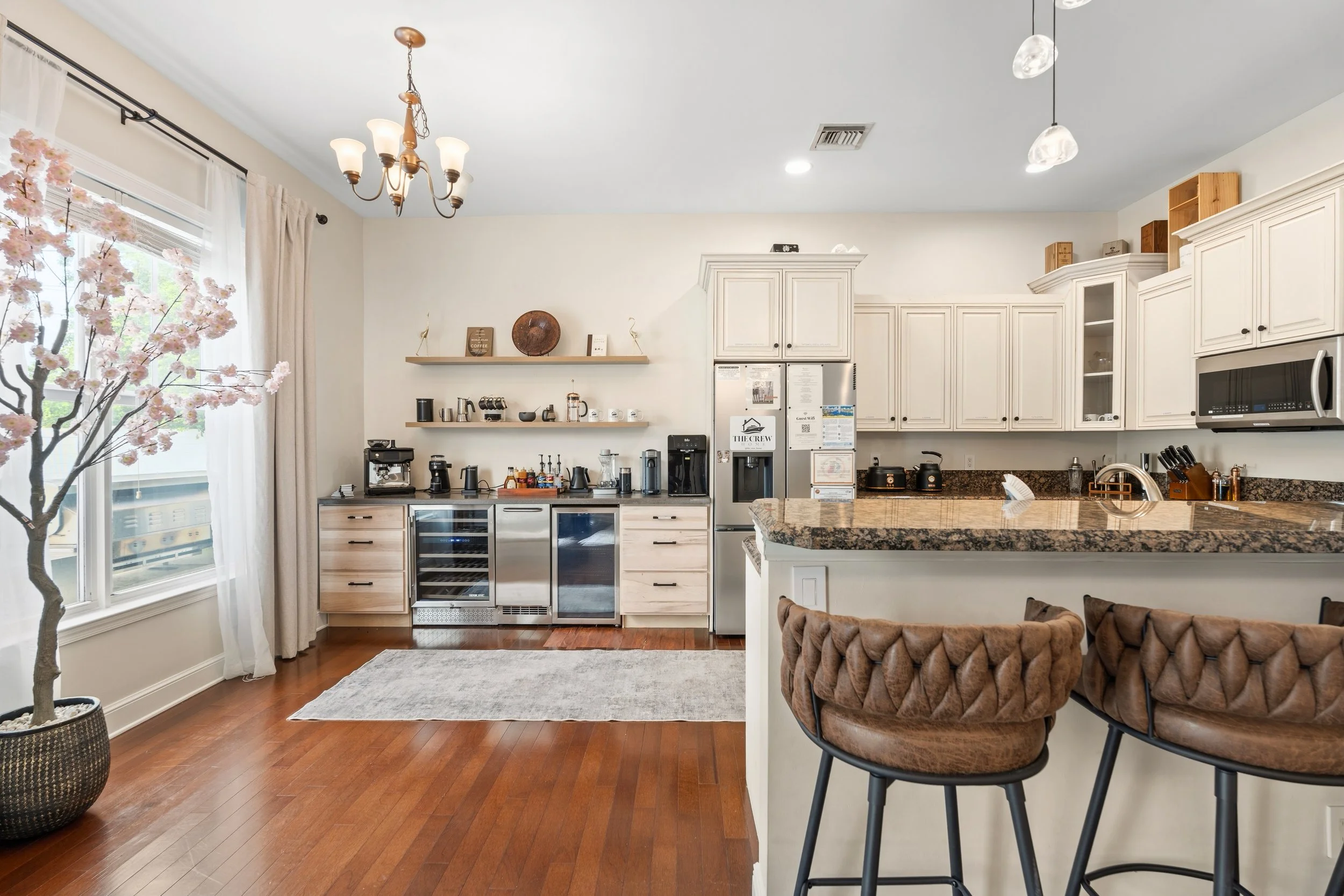 Kitchen area with white cabinets, granite countertop, coffee station, microwave, and bar stools, window with curtains, and cherry blossom tree in a pot.