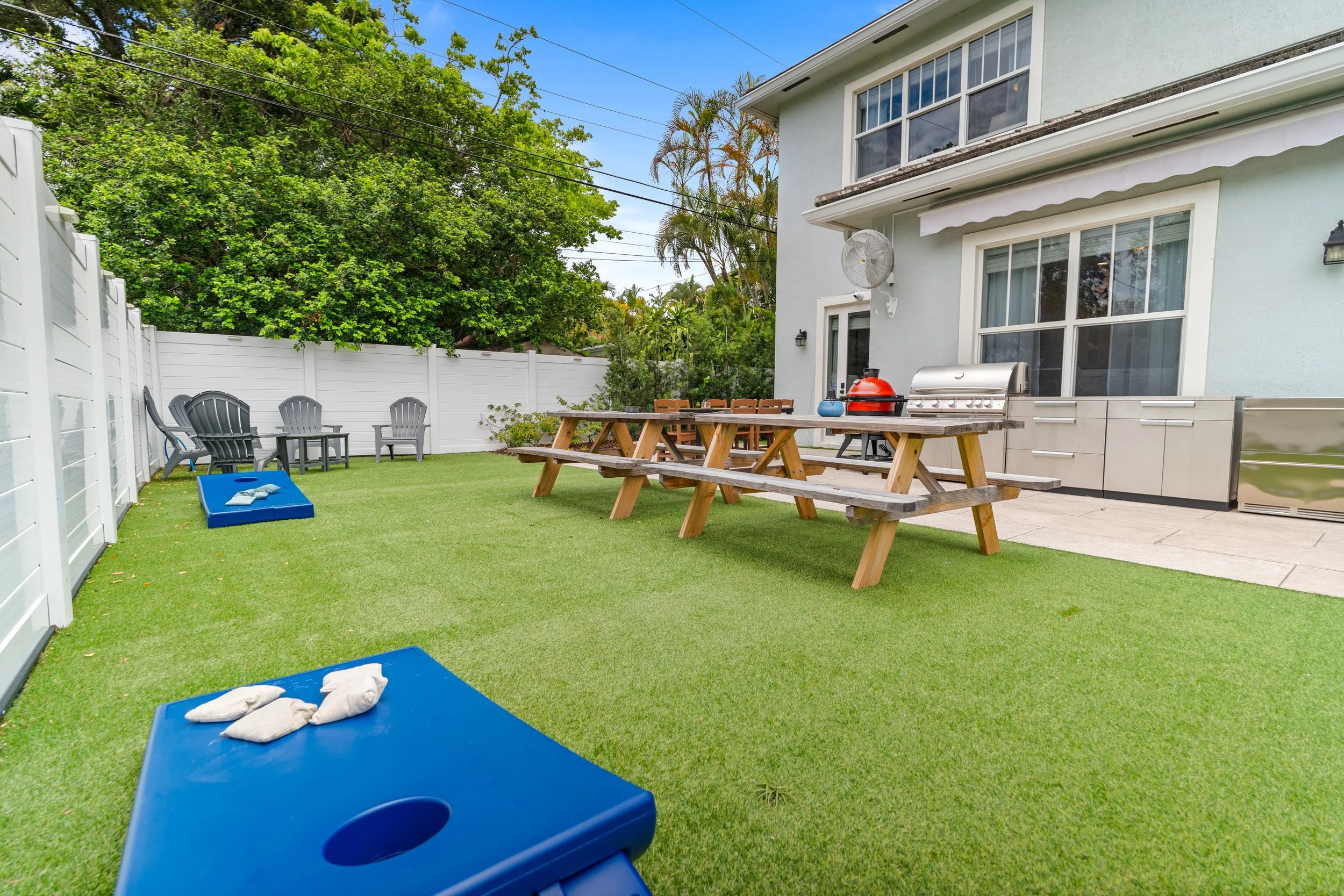 A backyard patio with green artificial grass, several gray outdoor chairs, a white fence, a large wooden picnic table with chairs, a gas grill, and a blue agility dog obstacle at the foreground. There are lush green trees and a clear blue sky in the 