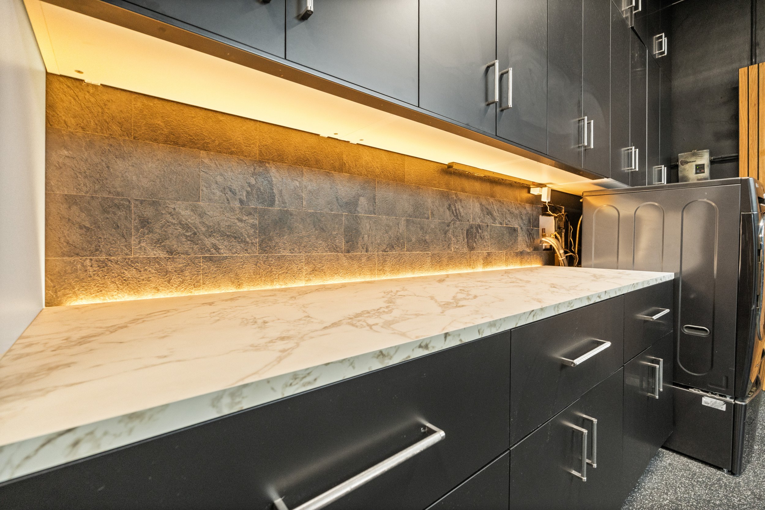 Close-up view of a modern kitchen countertop with dark cabinetry, a white marble surface, and a brown tile backsplash illuminated by warm lighting, with a washing machine at the side.