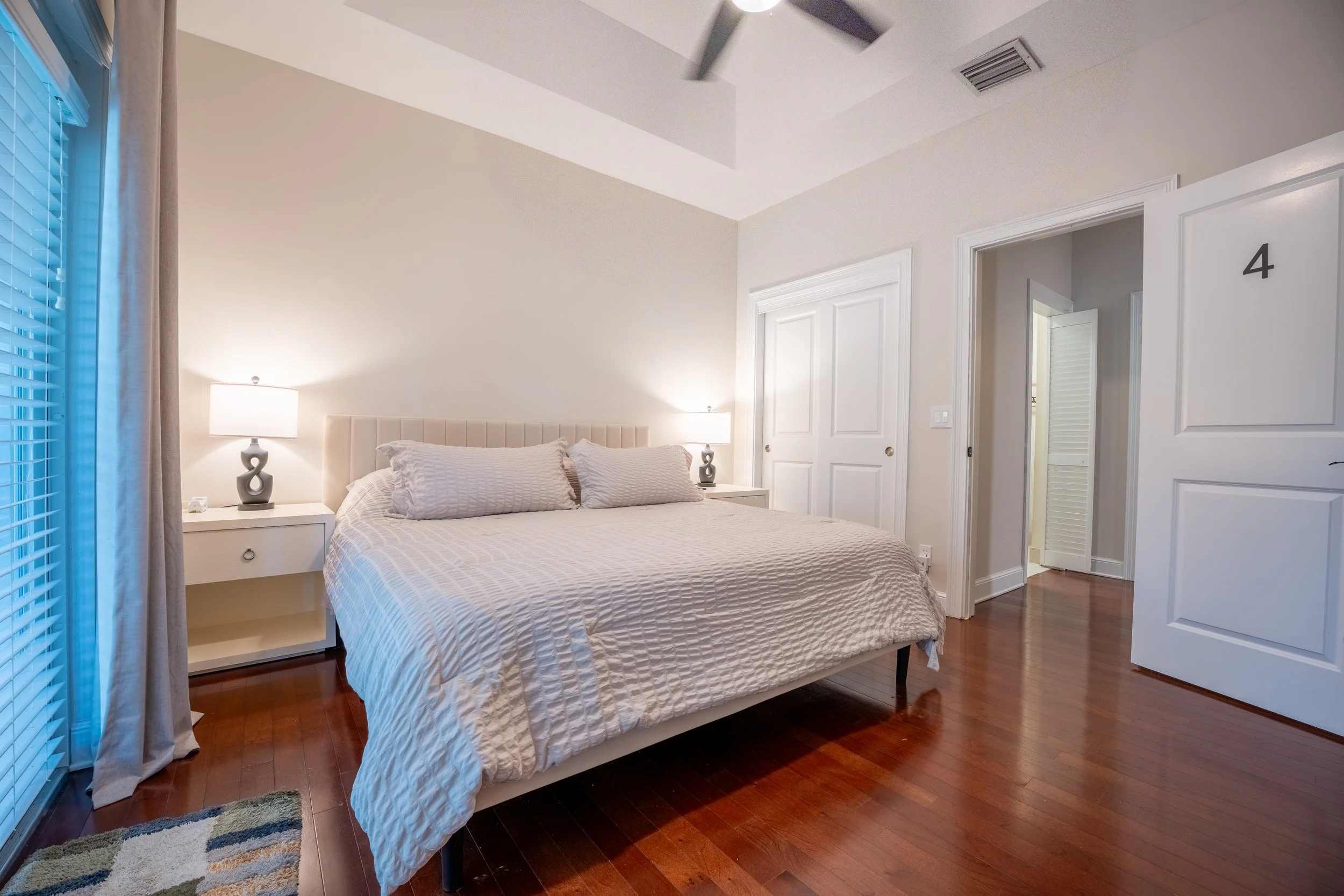 A bedroom with a bed, white bedding, two matching white nightstands with lamps, a window with blinds, and a door with the number 4 on it. The room has hardwood floors and white walls.