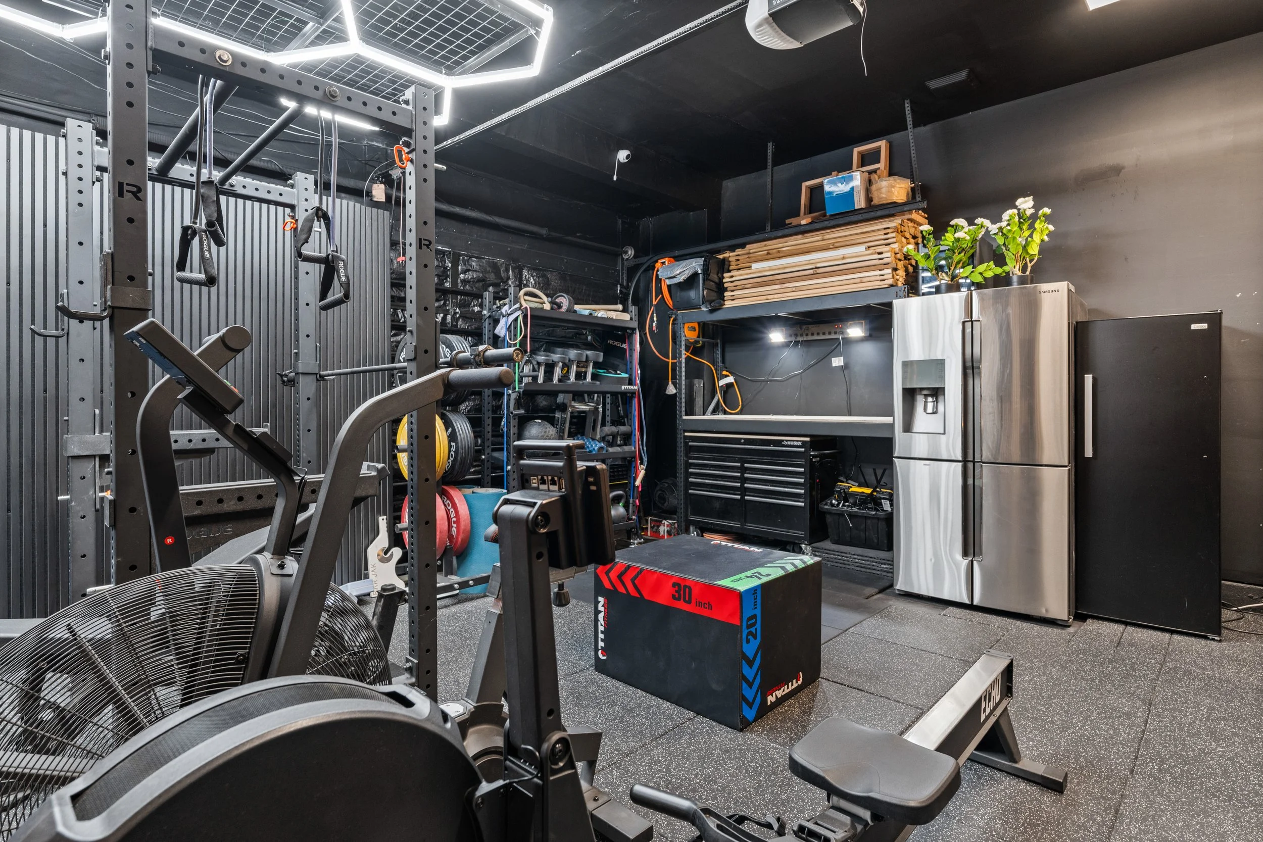 A gym with various fitness equipment, including a treadmill, weights, resistance bands, and a box for box jumps, with a storage shelf holding wood planks, a plant, and a refrigerator.