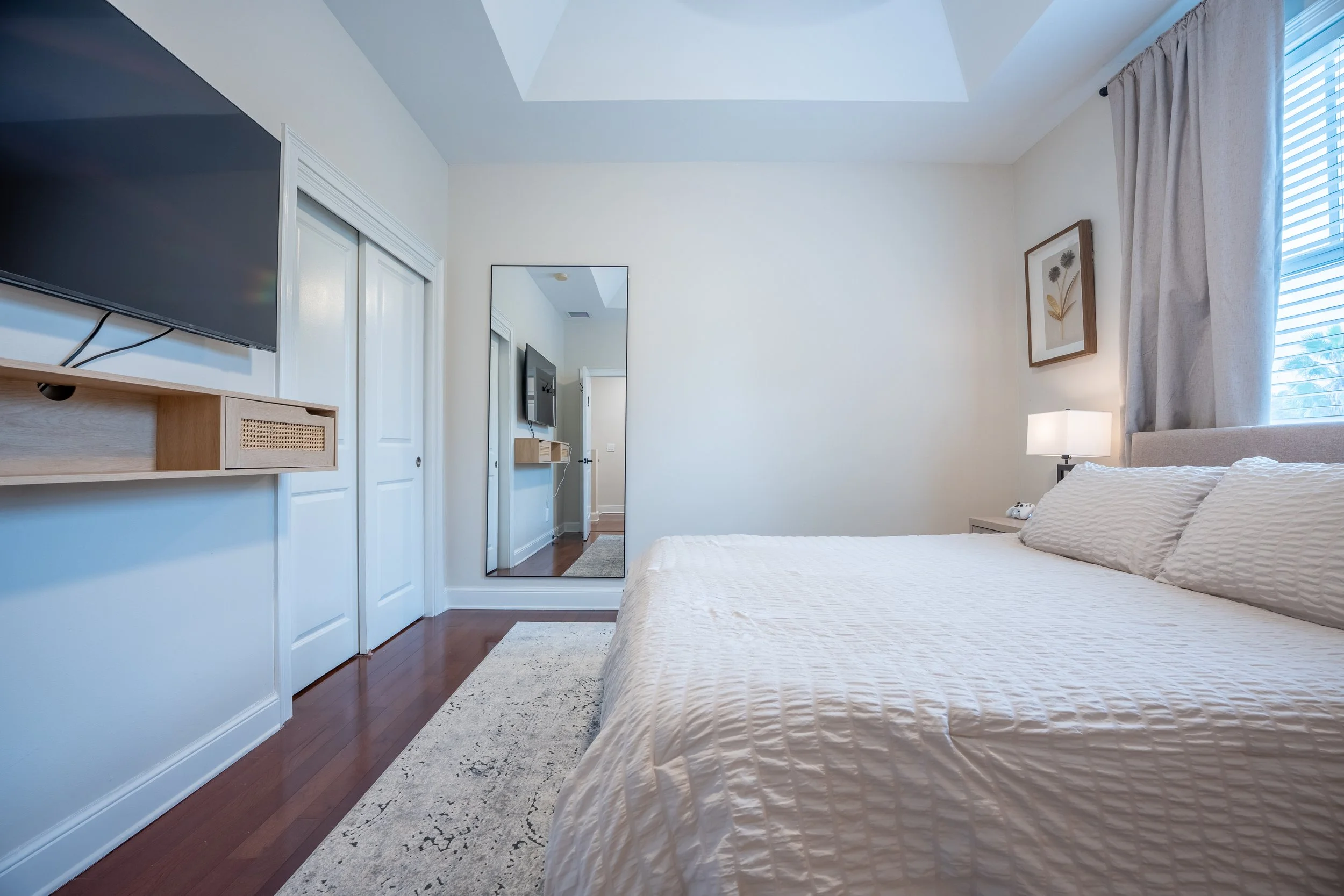 A bedroom with a bed, nightstand with lamp, wall art, large window with curtains, full-length mirror, wall-mounted TV, and hardwood floors.