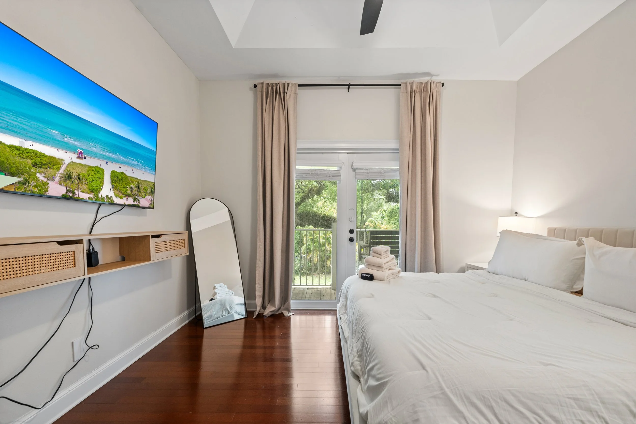 A bright bedroom with a large bed dressed in white linens, a sliding glass door with beige curtains leading to a balcony, a wall-mounted TV displaying a beach scene, a mirror leaning against the wall, and a small pile of white towels on a chair near 