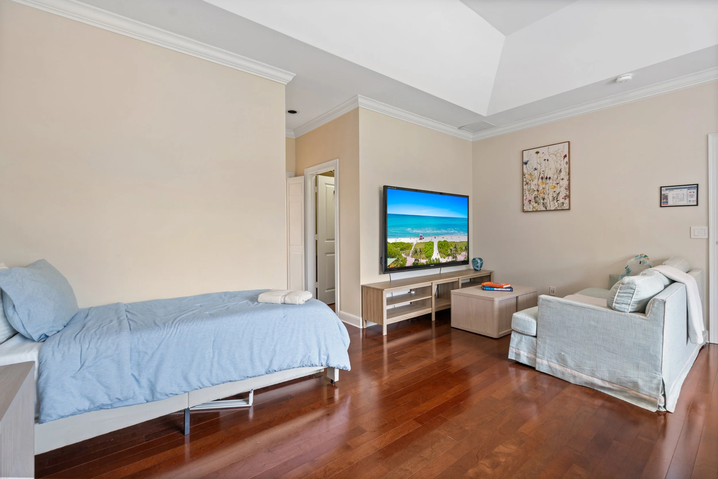 A bedroom with a bed covered in a light blue sheet, hardwood floors, a television on a wooden stand showing an ocean view, a cream-colored armchair, and wall art with floral designs.
