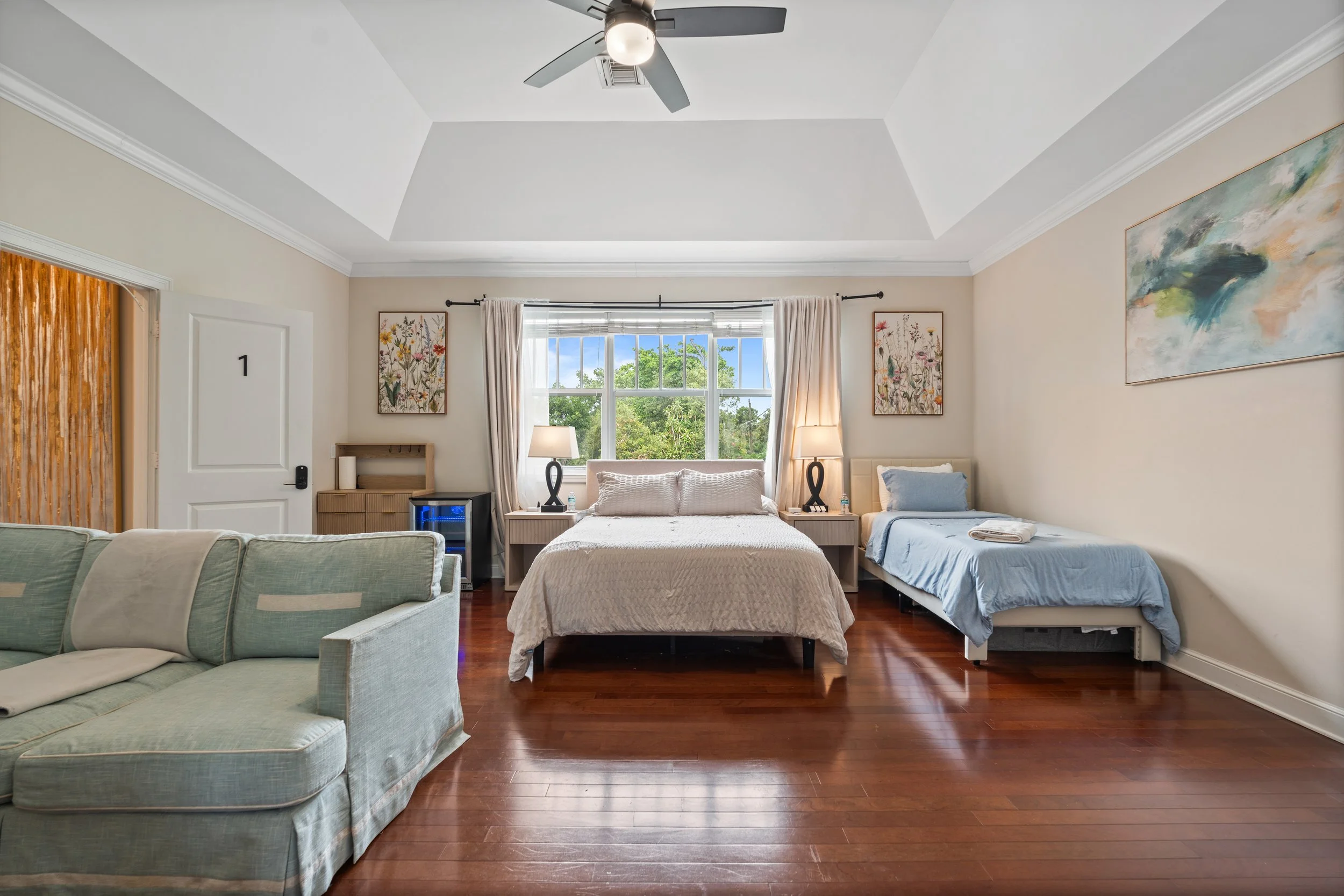 A bedroom with a queen-sized bed in the center, two bedside tables with lamps, a window with curtains, a painting on the right wall, and a ceiling fan. There is an additional single bed on the right side and a sofa in the foreground.