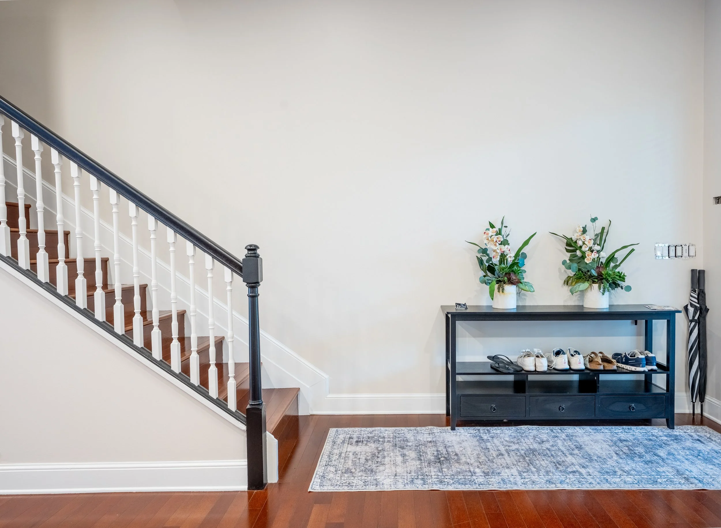 Indoor hallway with stairs, a dark wood console table with white flower arrangements, a patterned rug, and a shoe rack with various shoes.