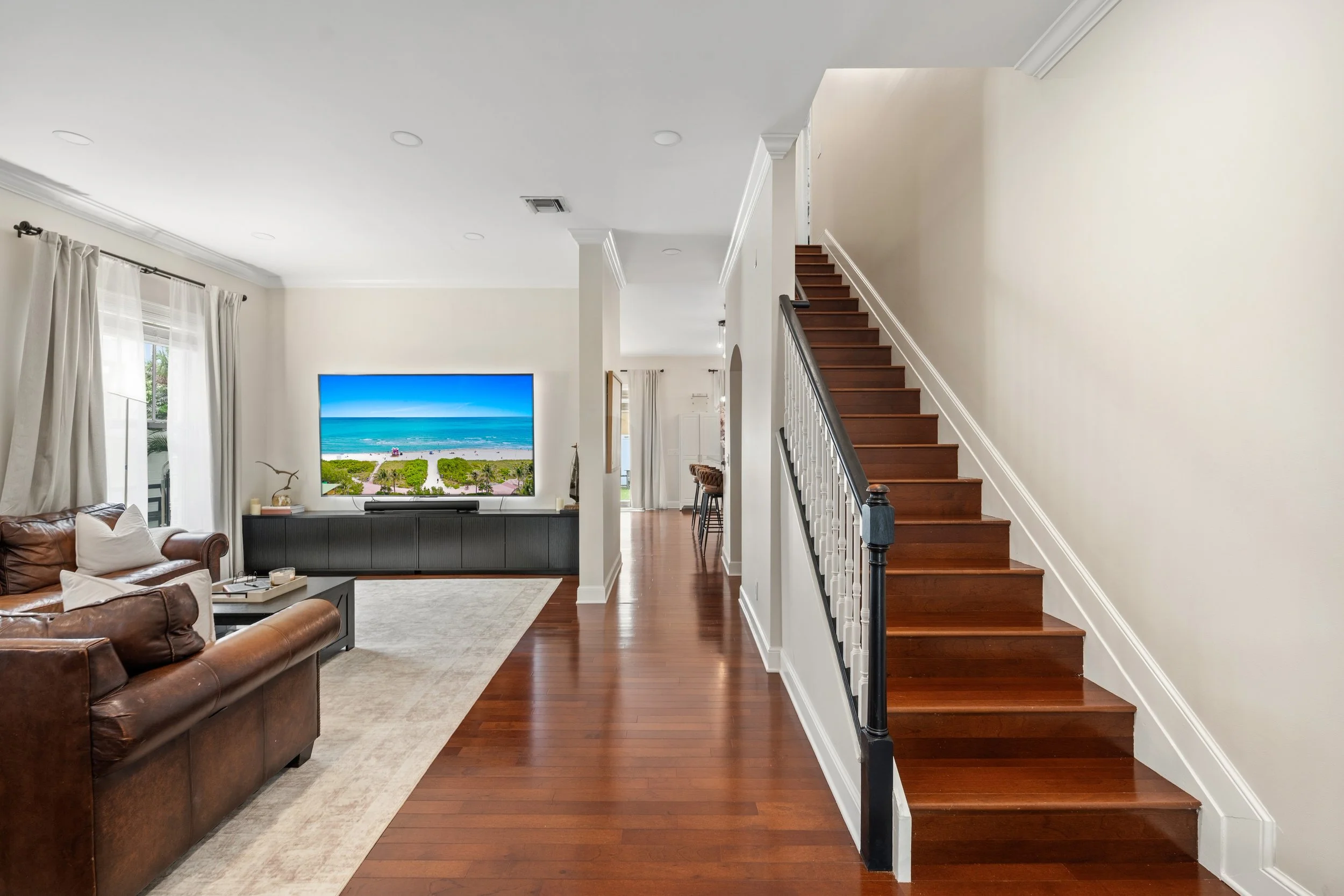 Living room with leather sofas, a large TV showing a beach scene, hardwood floors, and a staircase leading to the second floor.