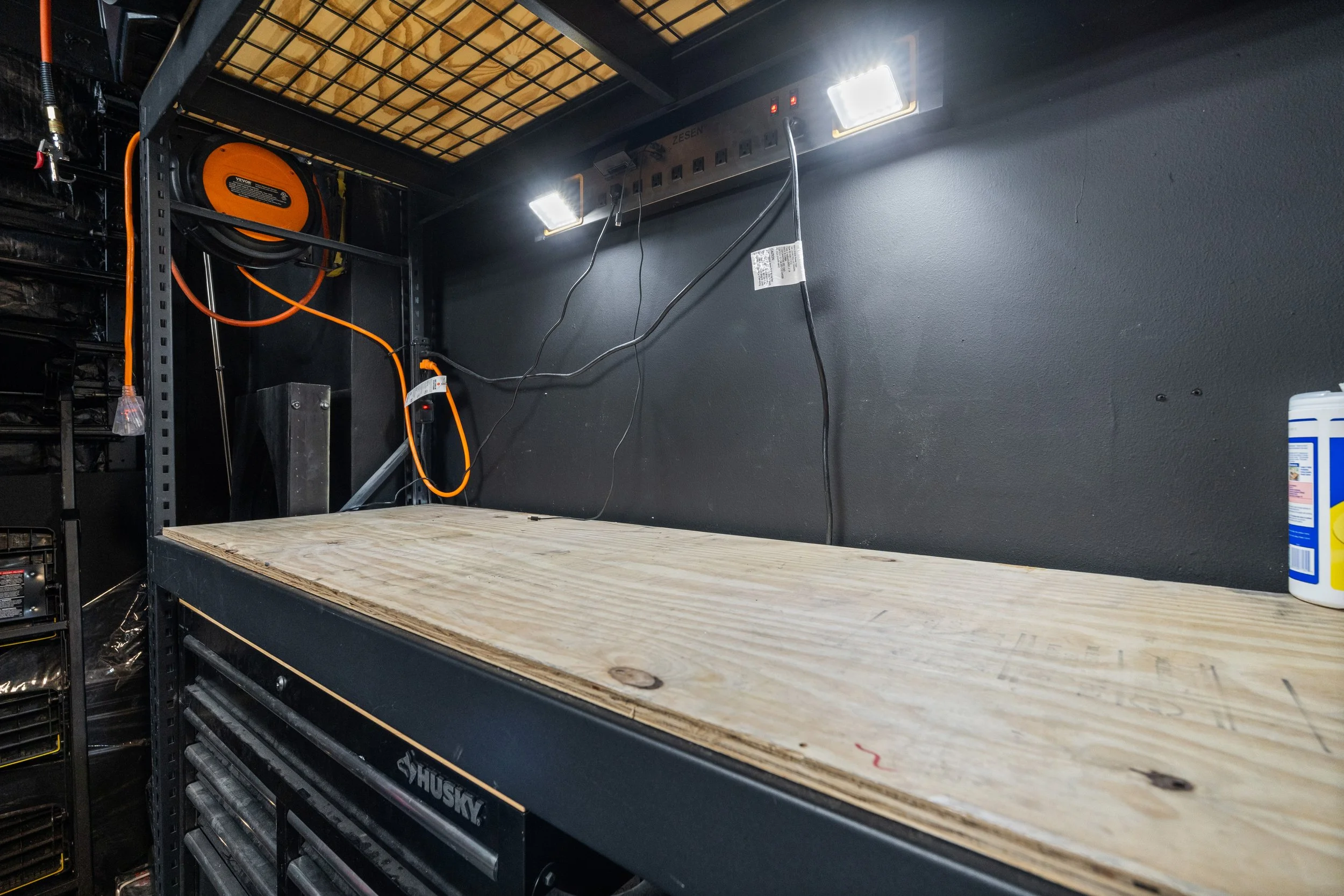 A workbench with a wooden top in a workshop, with electrical wiring and a power strip mounted above, and a container of disinfectant wipes on the right side.