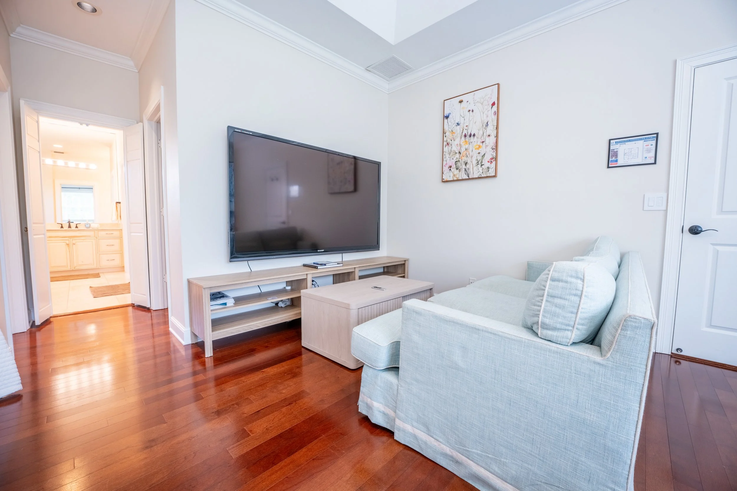 Living room with a beige couch, wall-mounted flat-screen TV, wooden entertainment stand, framed floral artwork, and hardwood floors.