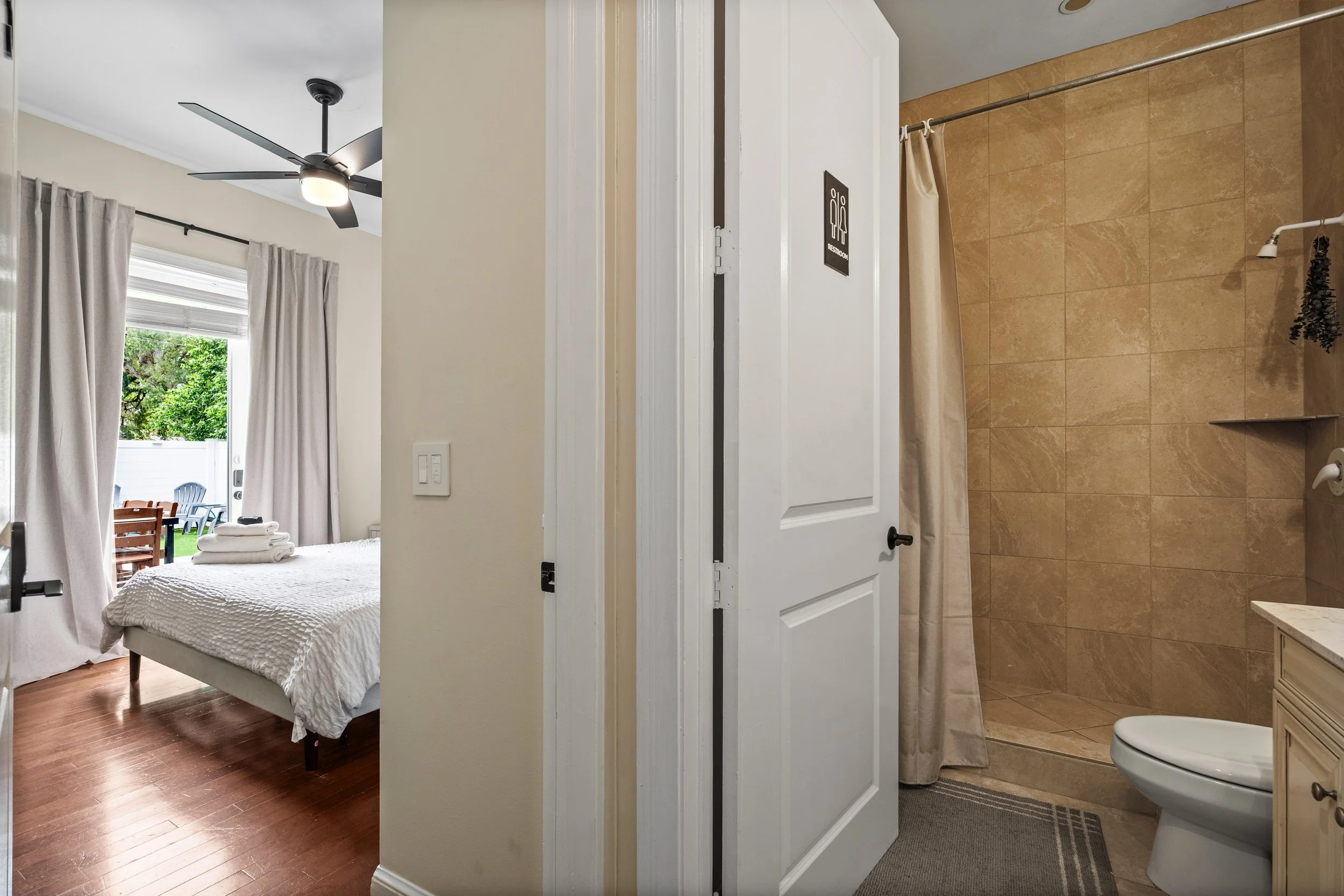 A bedroom with a bed, dresser, and window with curtains, visible through an open door from a bathroom with tile flooring, shower curtain, and toilet.