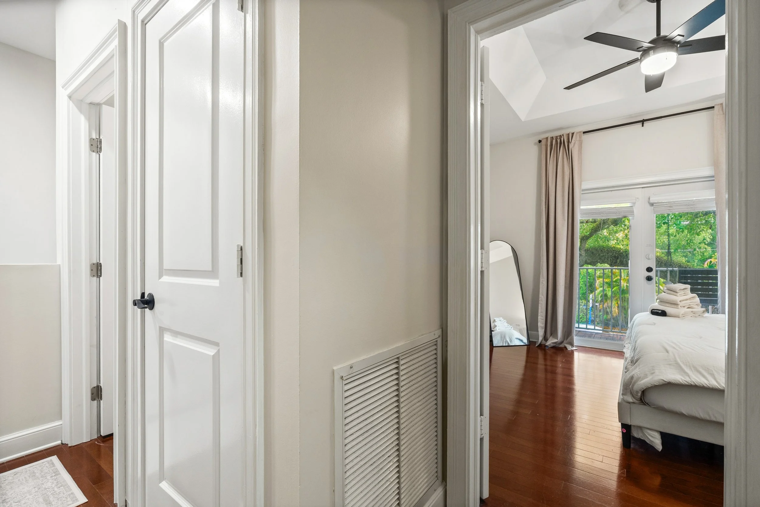 View from hallway into bedroom with sliding glass door, trees outside, bed with stacked towels, ceiling fan, and beige curtains.
