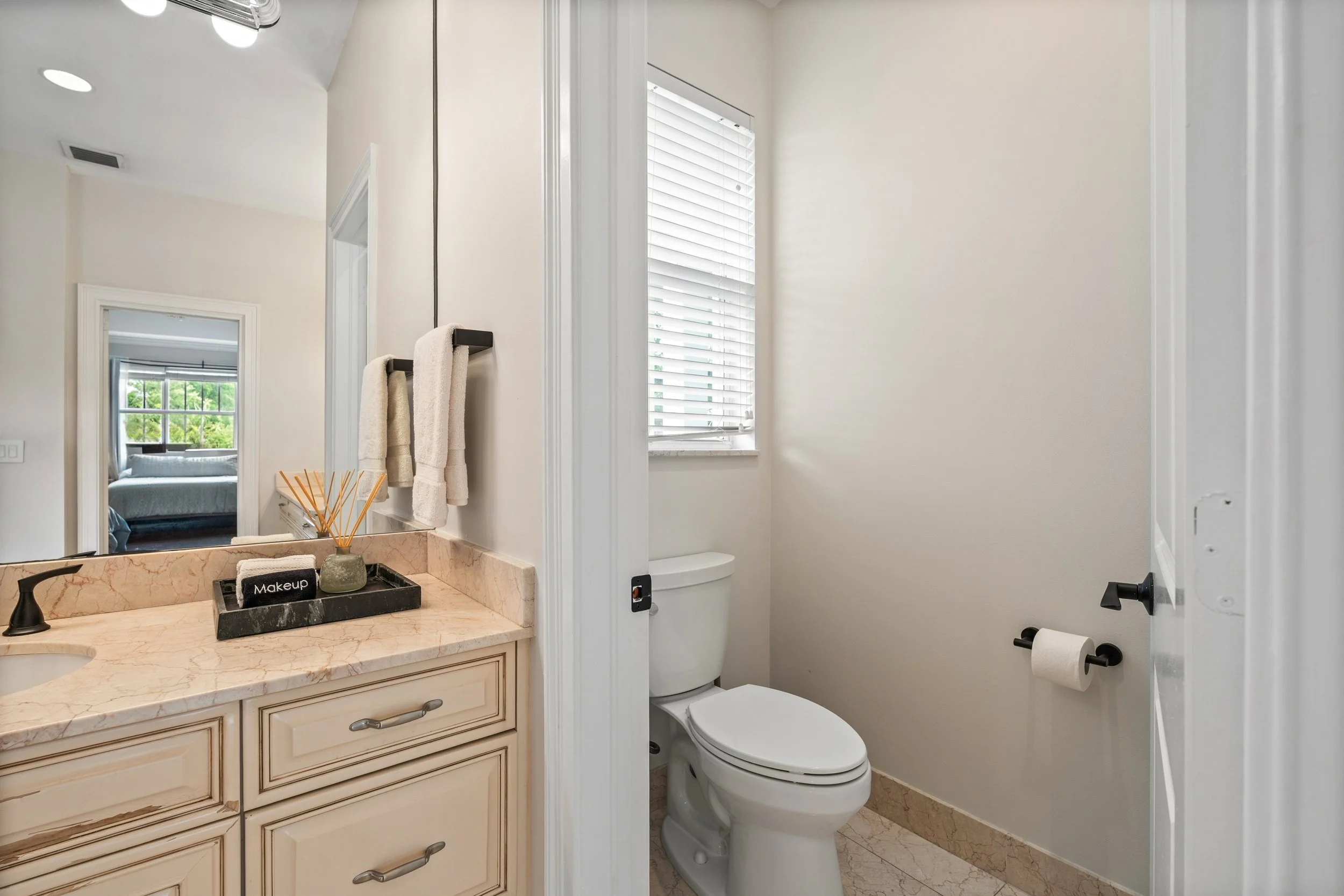 A small bathroom with a vanity countertop, mirror, and a toilet. The vanity has a marble surface with a black tray containing a makeup brush, and towels hanging on a black towel bar. There is a window with blinds allowing natural light. The room has 
