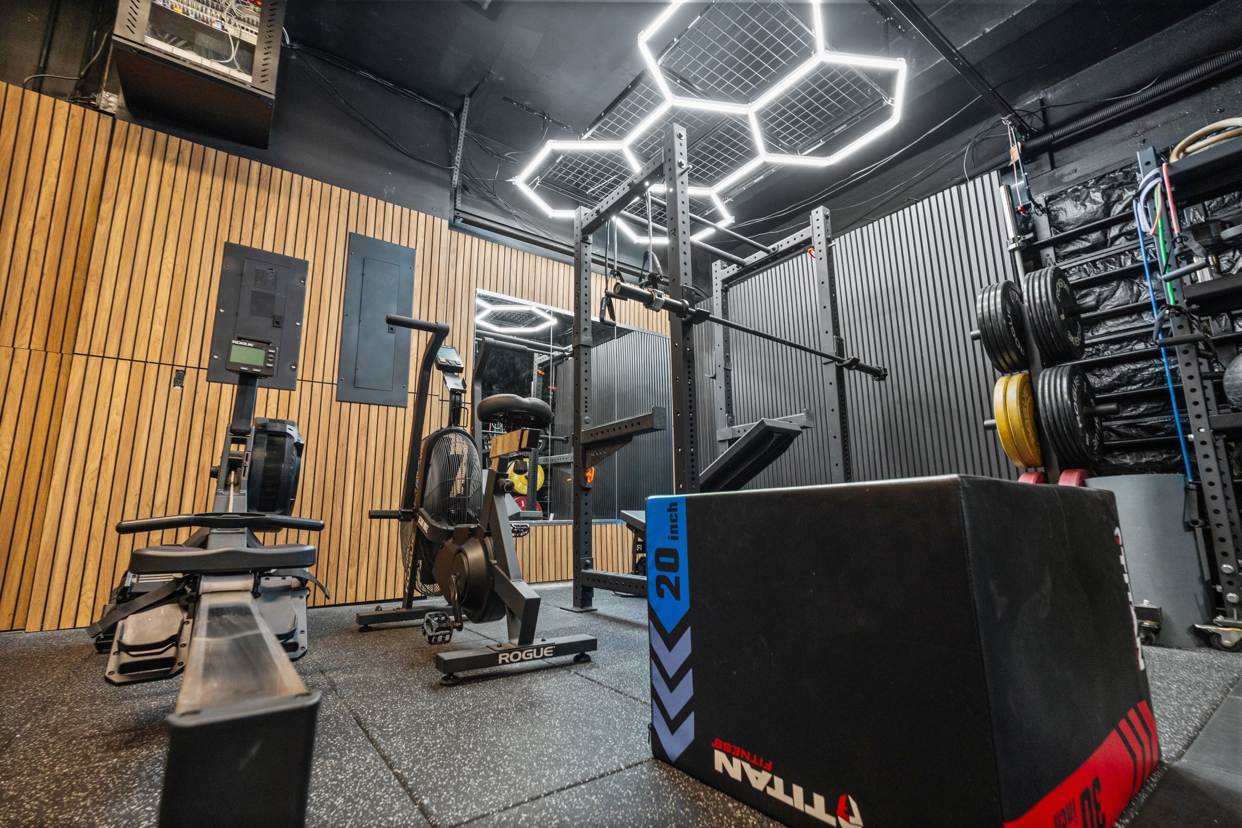 A small gym with black walls and a wooden wall panel. The ceiling has hexagon-shaped LED lights. There is workout equipment including a rowing machine, a squat rack with a barbell, and weight plates on a rack. There is also a black plyometric box wit
