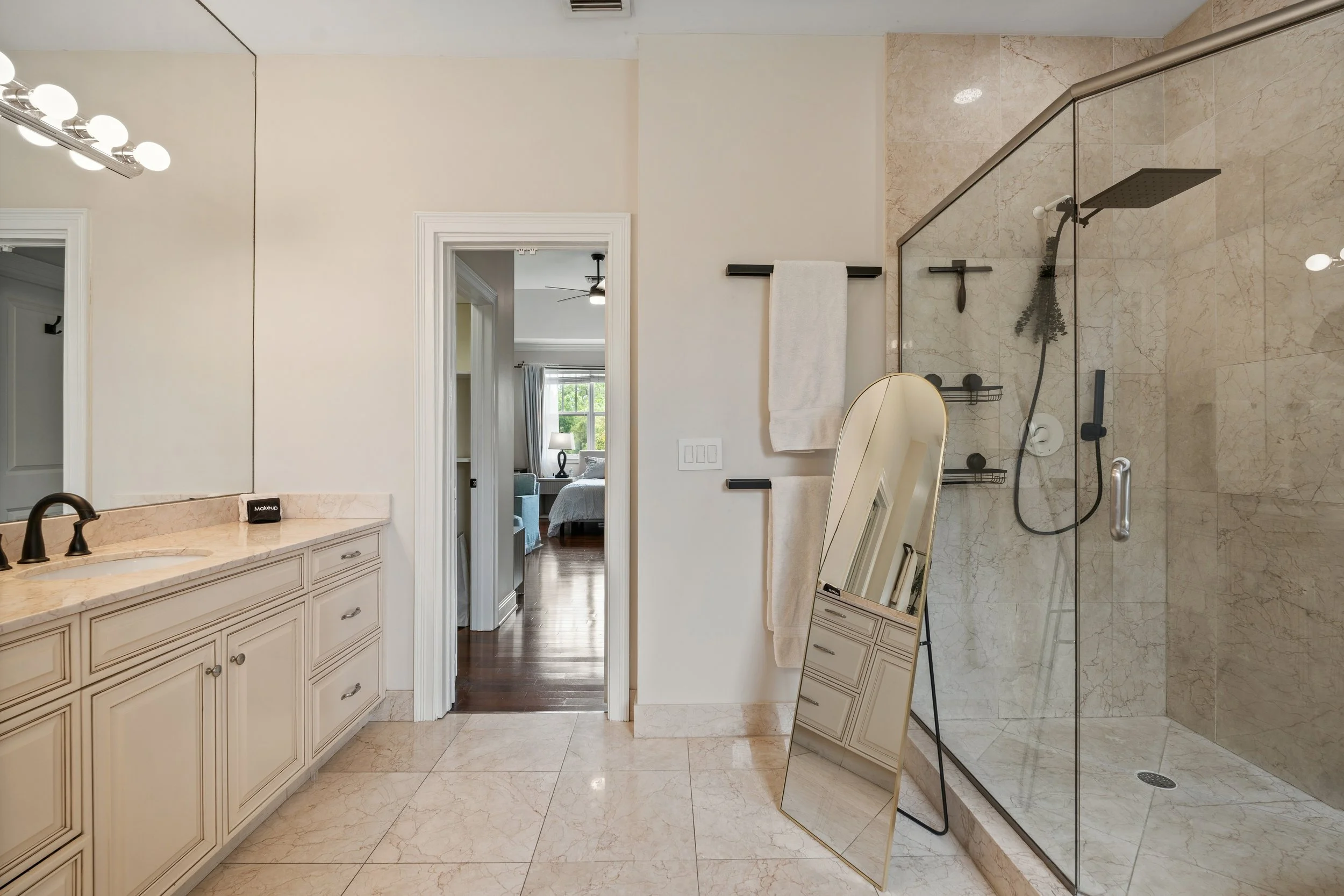 Bathroom with a glass shower enclosure, beige marble tiles, white cabinetry, and a doorway leading to a bedroom with a bed, nightstand, and window.