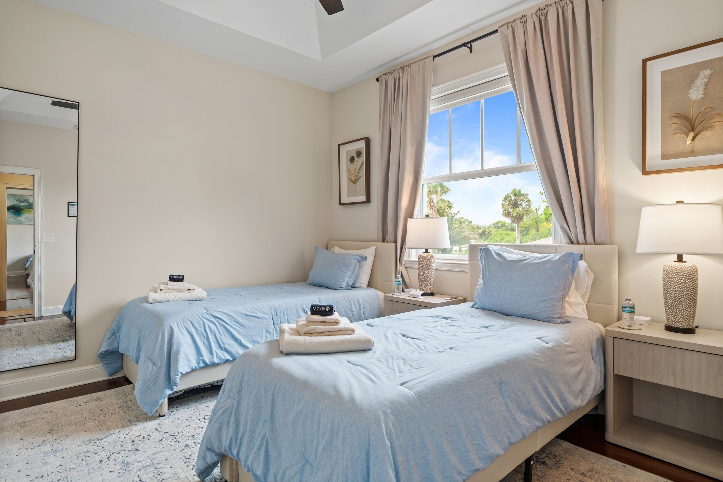 A bedroom with two twin beds, each with light blue bedding, pillows, and a stack of towels with a black makeup bag on top. There are two nightstands with lamps and water bottles. A window with beige curtains shows trees and a blue sky outside. Wall a