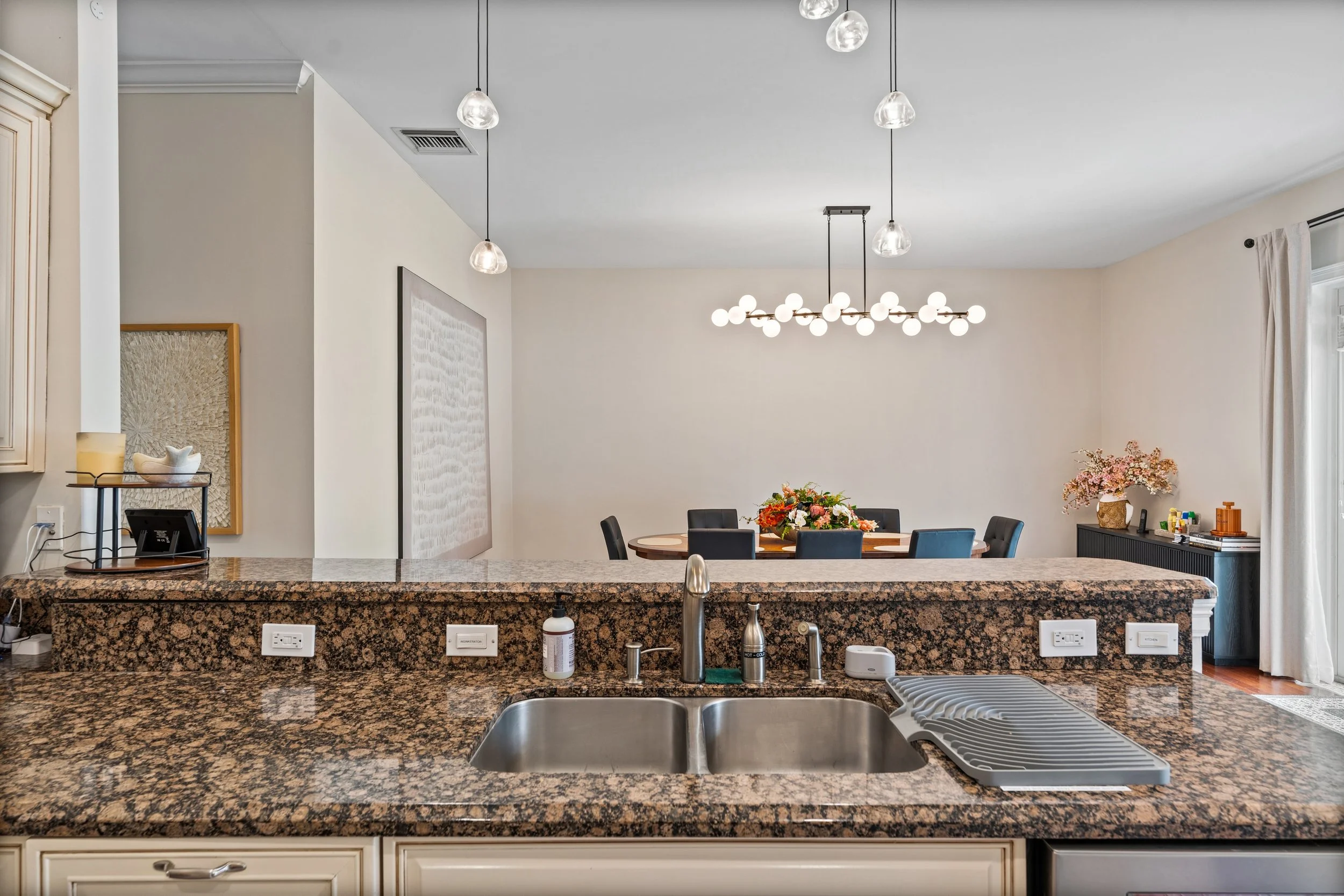 View of a kitchen with a granite countertop, sink, and faucet in the foreground. In the background, there is a dining area with a round table, chairs, and a floral centerpiece. Modern pendant lights hang from the ceiling over the dining table. The wa