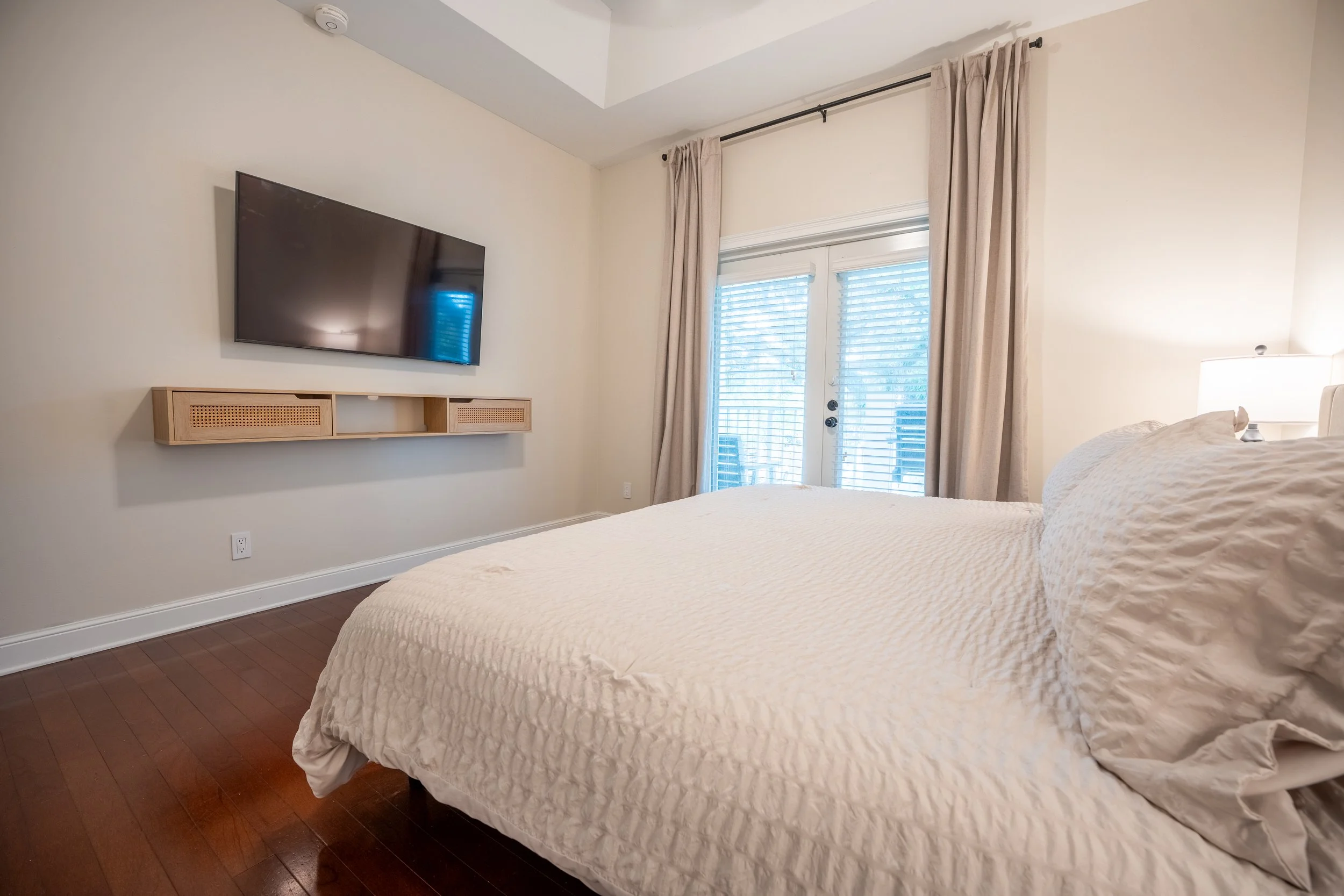 A bedroom with a bed covered in a beige textured quilt, a bedside table with a lamp, a wall-mounted TV above a wooden shelf, and large windows with blinds and beige curtains.
