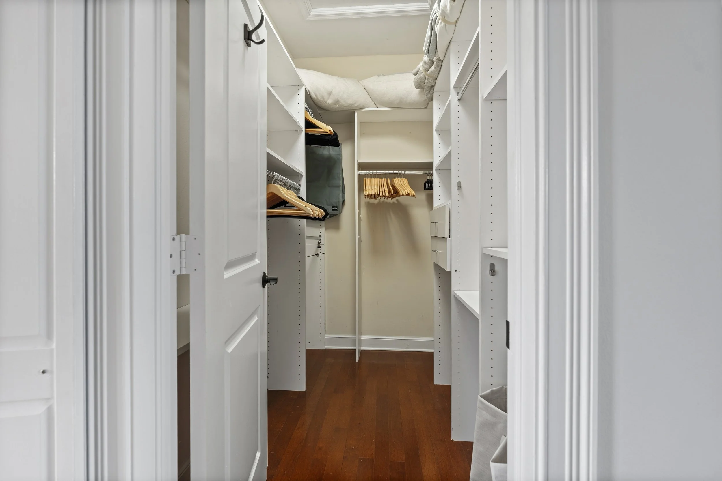 Empty walk-in closet with white shelves, hanging rods, and a bed with pillows on a slanted ceiling in the background.