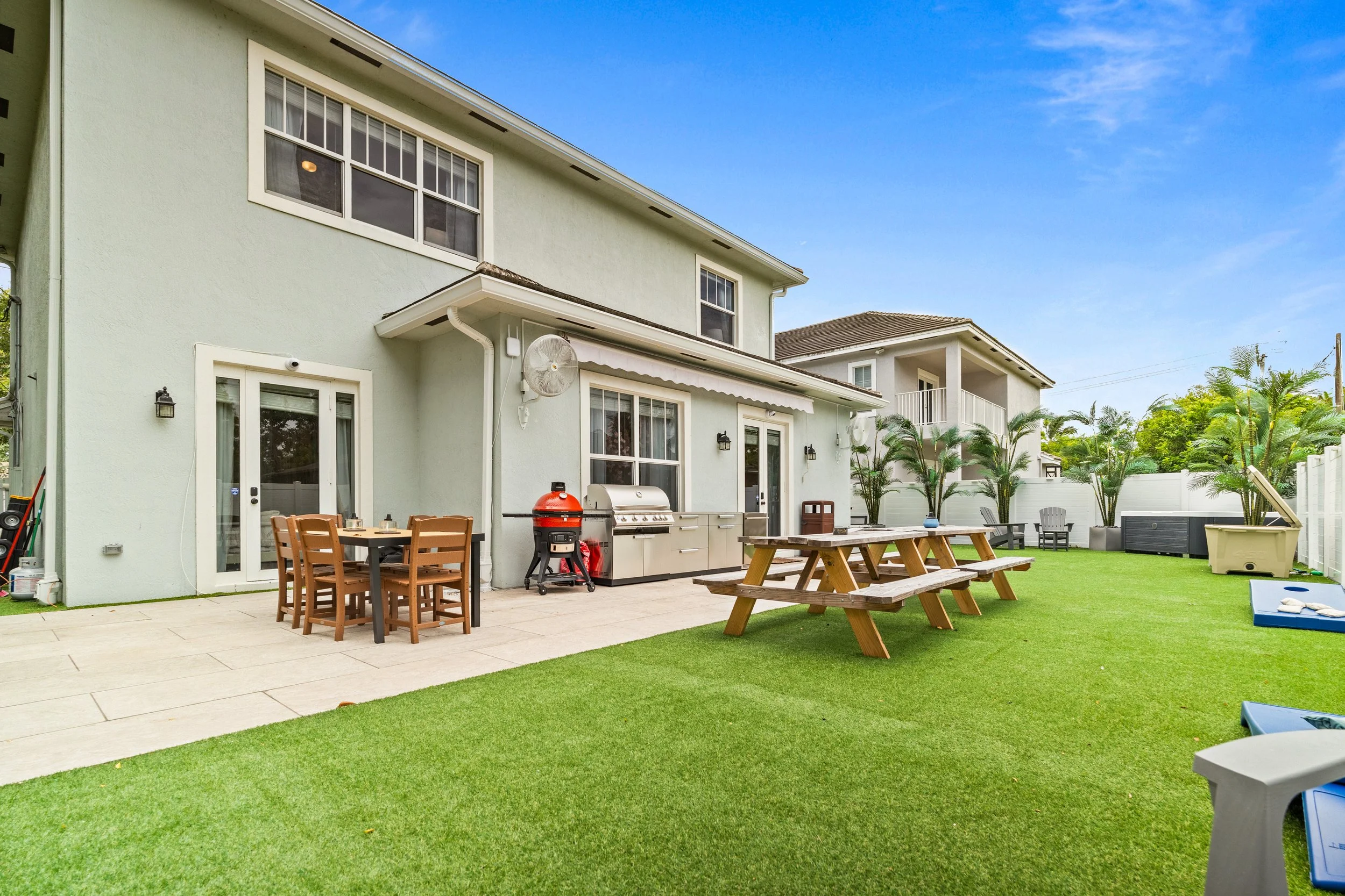 Backyard patio with outdoor seating, dining table, barbecue grill, and lush green lawn with trees and houses in the background under a clear blue sky.