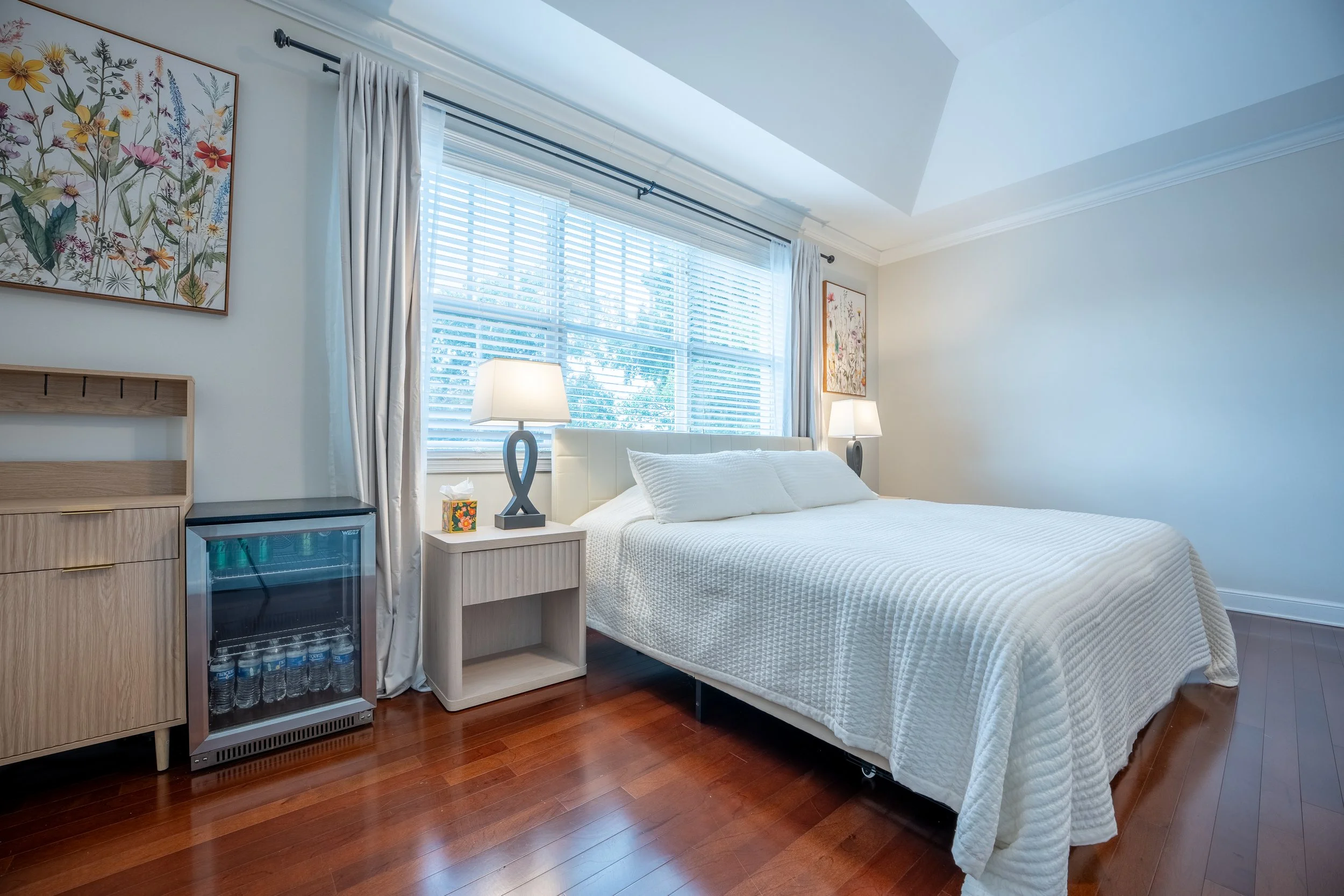 A bedroom with a bed, bedside tables with lamps, window with white blinds and curtains, wall art, and a mini fridge filled with bottled water.