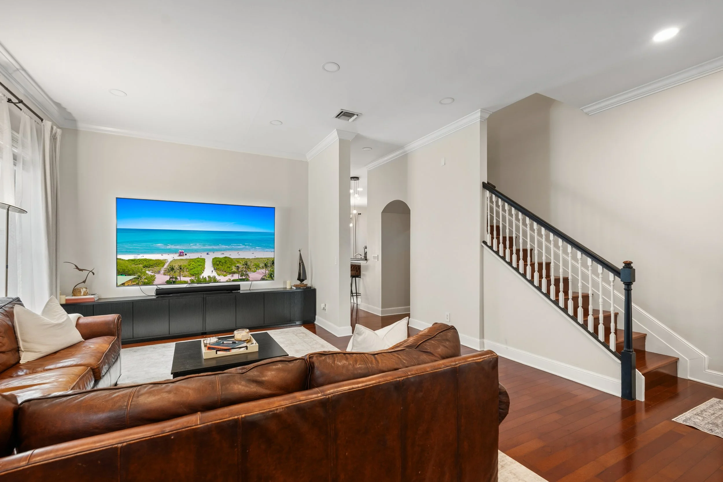 Living room with a large flat-screen TV displaying a beach scene, a brown leather sofa, a coffee table, and white curtains on the windows. There's a staircase with a black and white railing on the right.