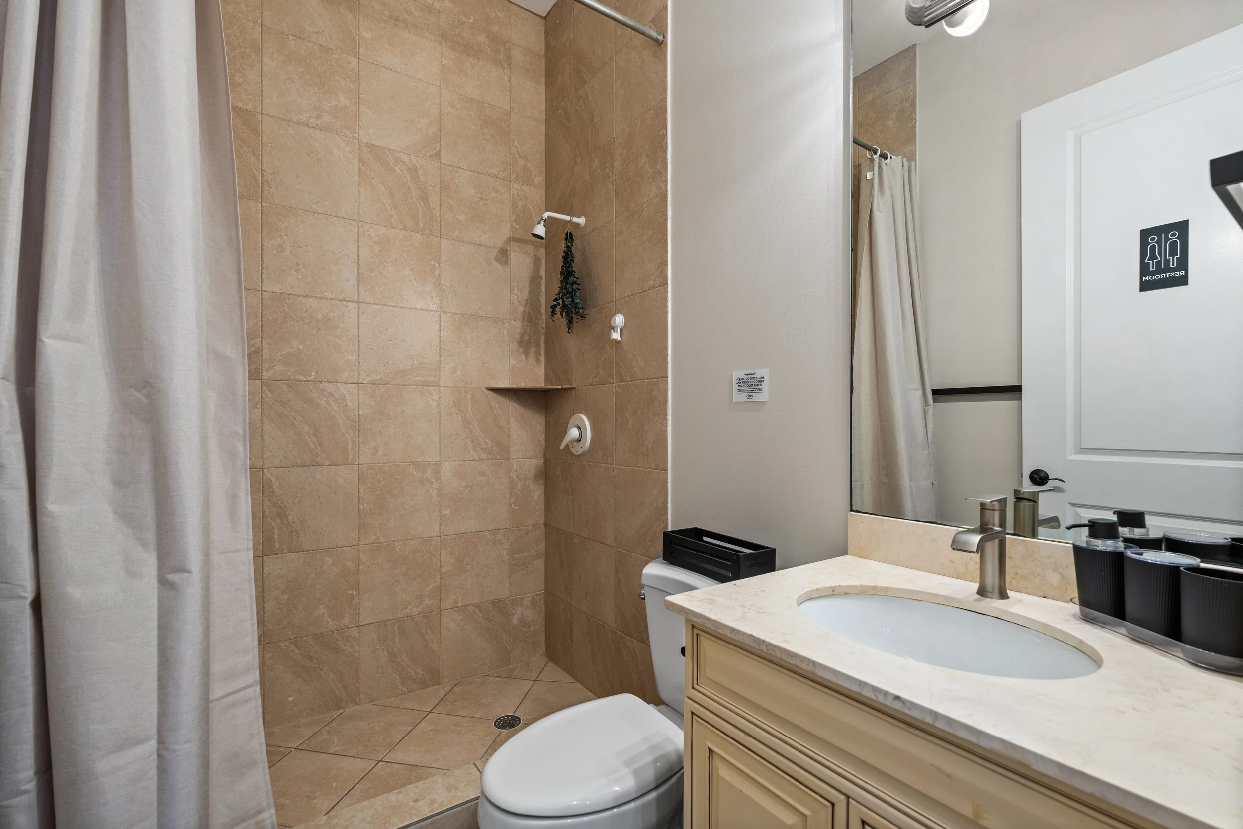 A bathroom with beige tile walls and floor, a shower area with a white shower rod and a hanging plant, a white toilet, a beige vanity with a marble countertop, a sink with a silver faucet, a large mirror, a soap dispenser, and a sign indicating the r