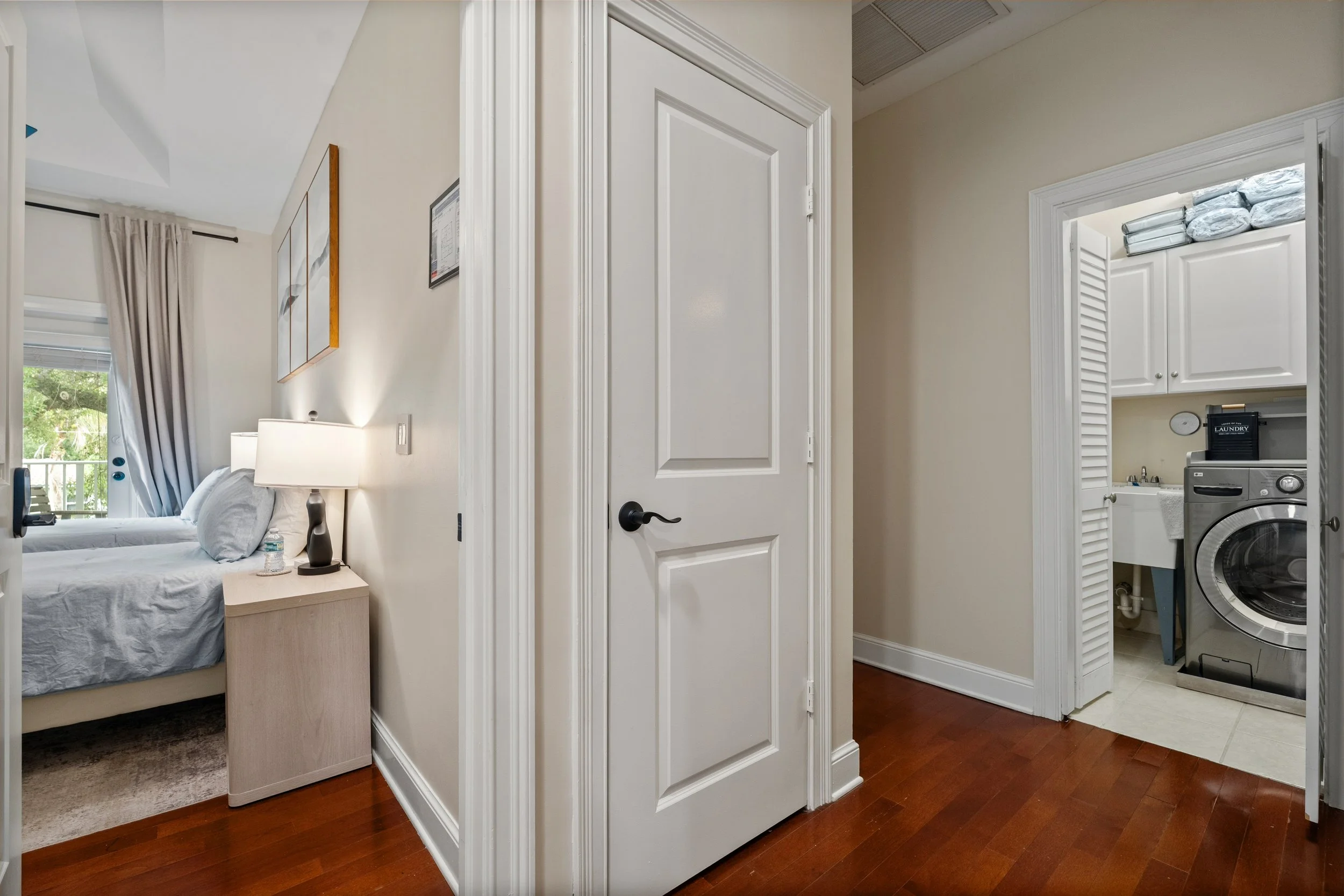 Interior view of a bedroom with a bedside table, lamp, and a bed with pillows. Part of a laundry area with a washing machine, cabinets, and laundry supplies is visible through an open door.