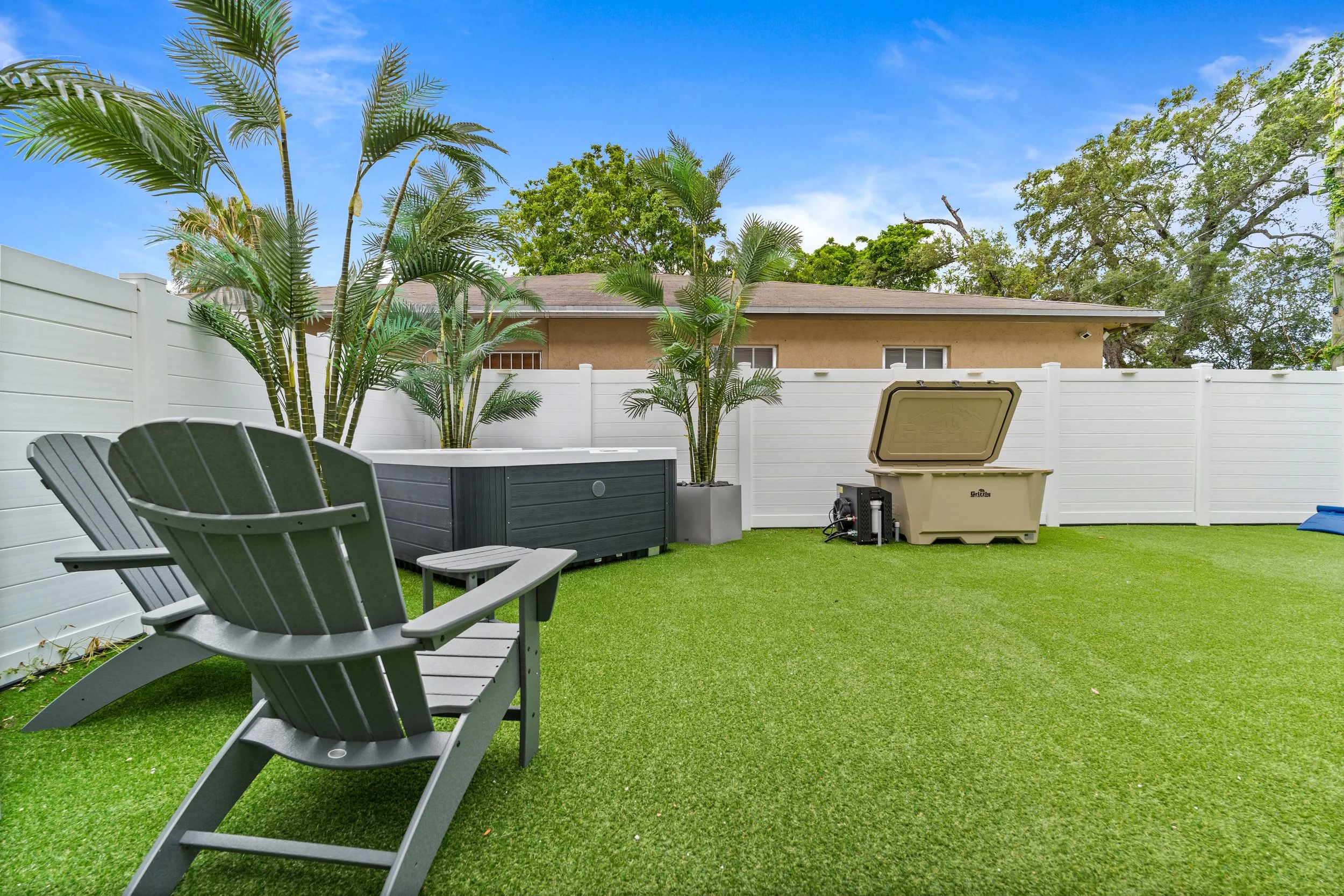 A backyard with green artificial grass, a white privacy fence, tropical plants, outdoor chairs, a hot tub, and a storage container, under a clear blue sky.