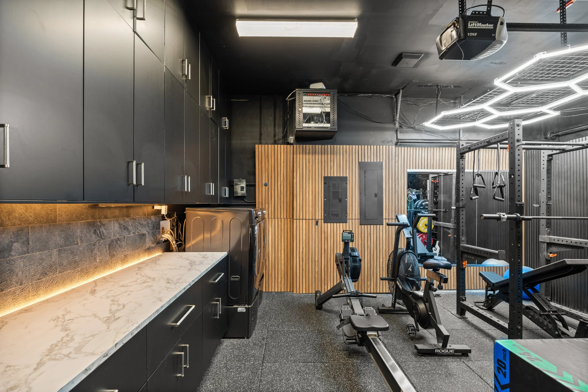 A modern gym with black cabinets, a marble countertop, and workout equipment including a rowing machine, exercise bike, and weight rack, with wood-paneled wall and hexagonal LED lights on the ceiling.