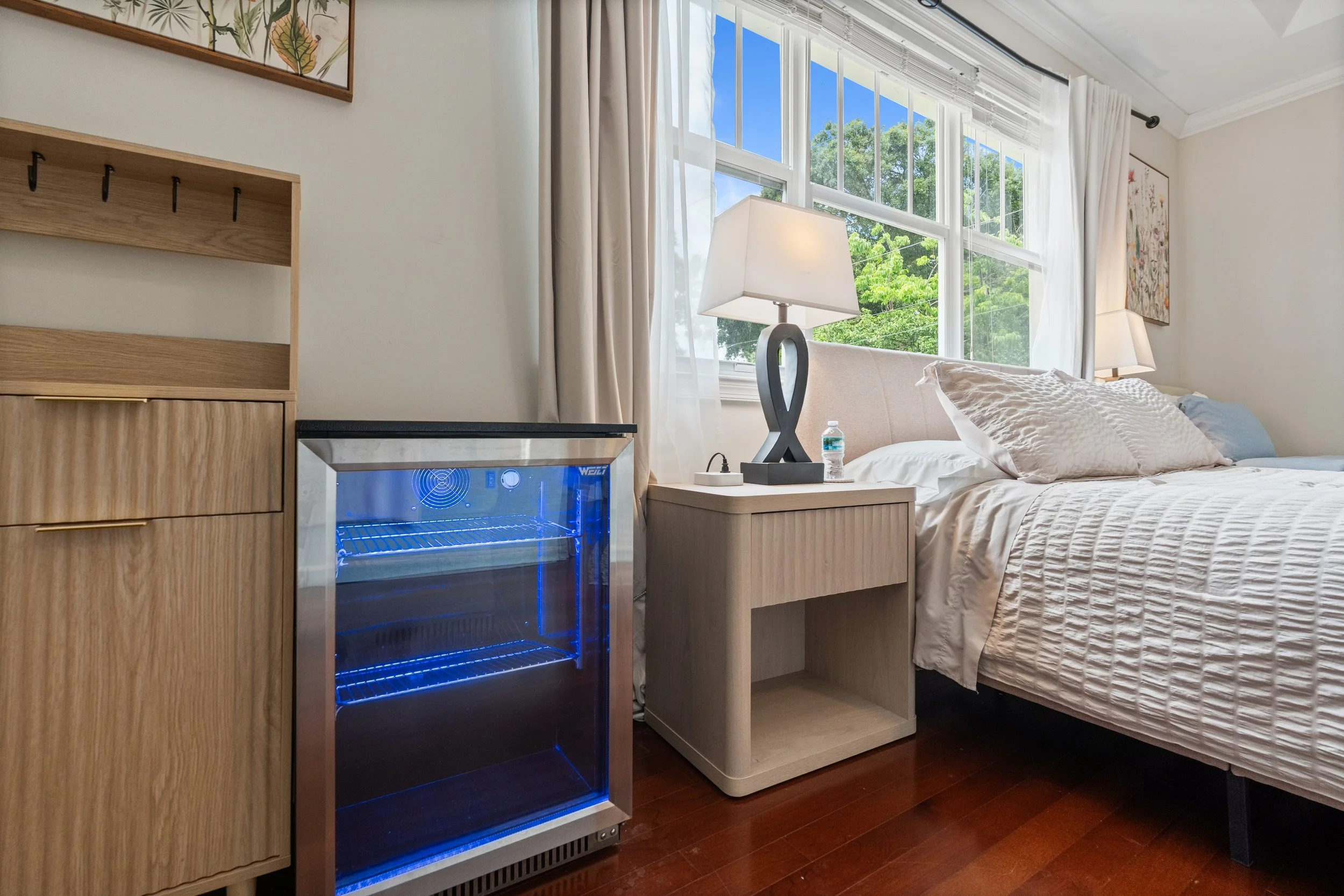 A bedroom with a bed, two nightstands with lamps, a window with white curtains, a small wooden cabinet, and a mini refrigerator with a blue interior light, all on a wooden floor.