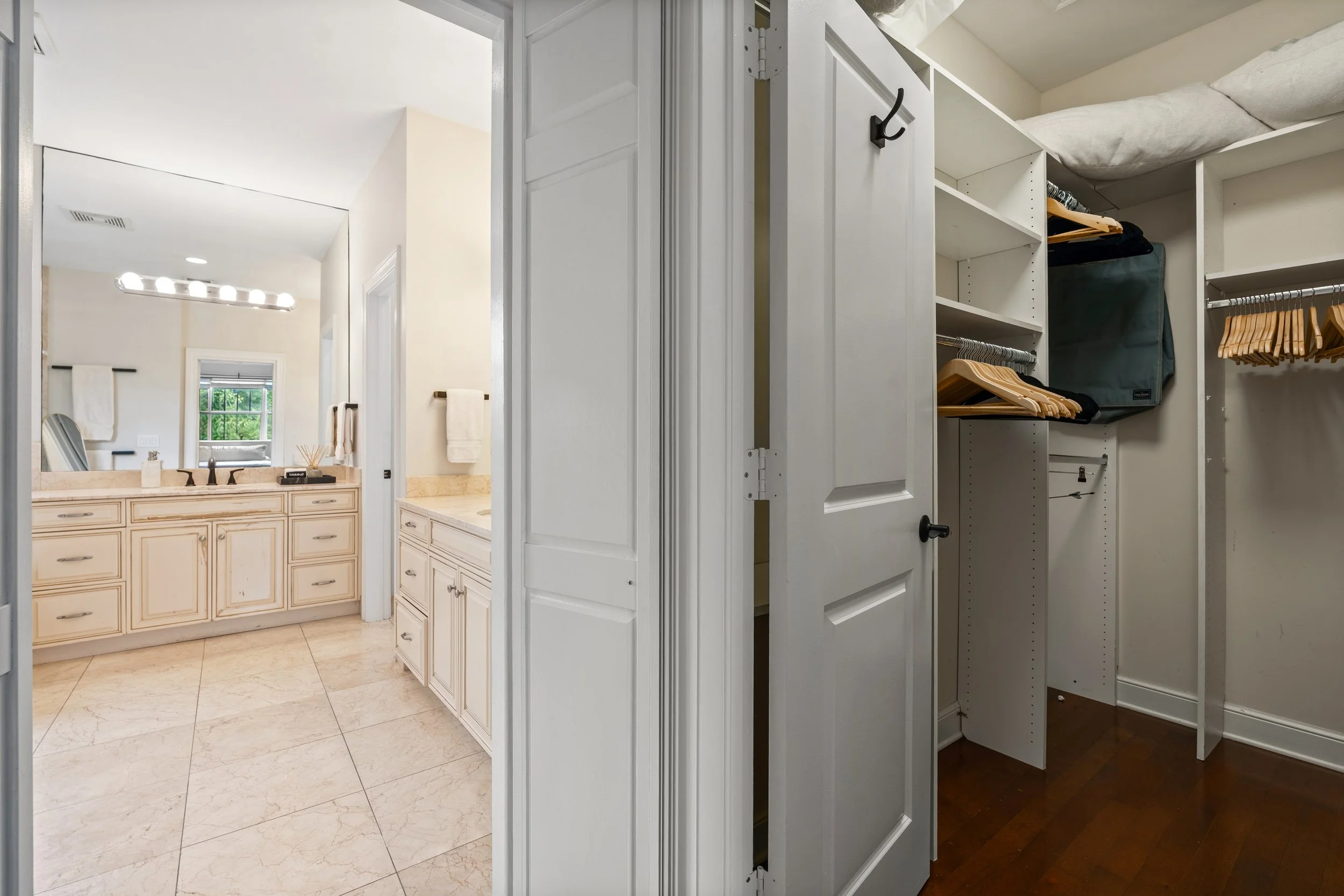 A view of a bathroom mirror and vanity on the left, and a closet with hanging rods and shelves on the right. The bathroom has cream-colored cabinetry and beige marble flooring. The closet contains light wood hangers, a bag, and pillows.