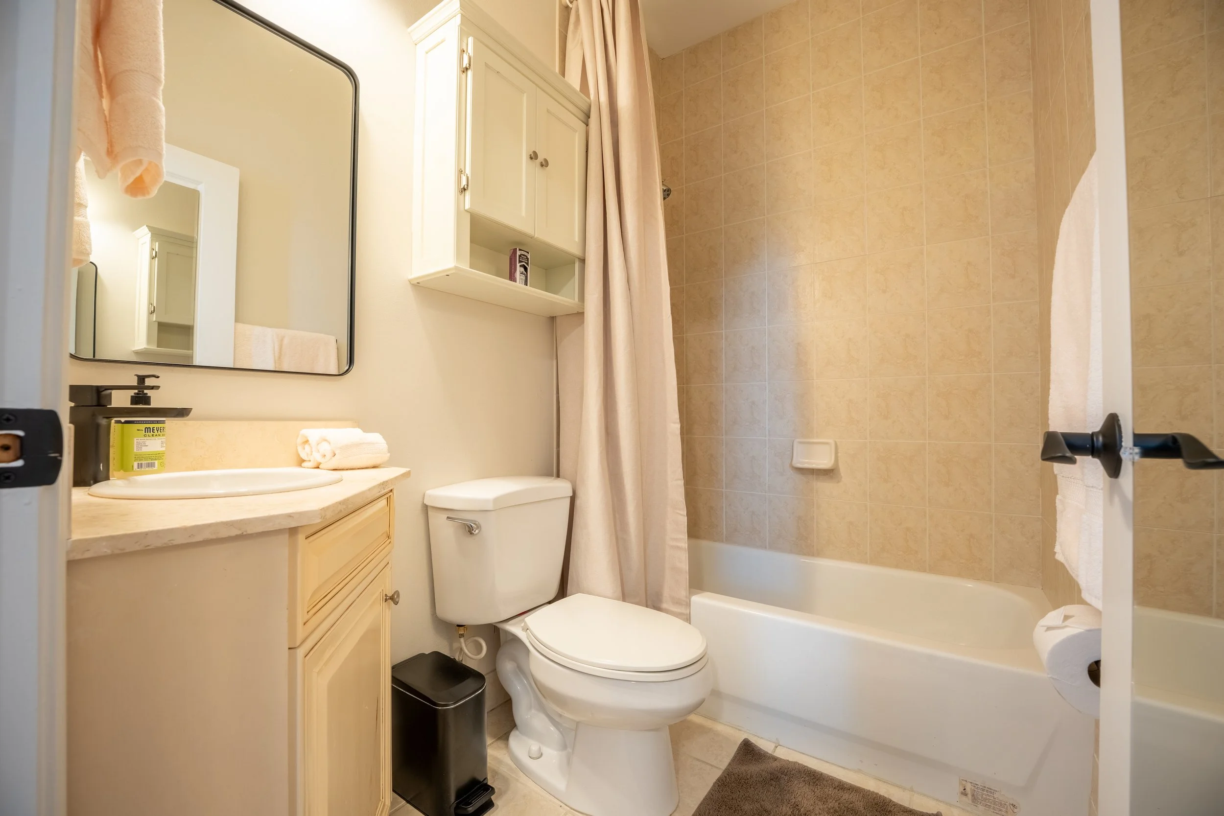 View of a small bathroom featuring a shower with beige tiles, a white toilet, a beige vanity with a mirror, a hand towel, a small cabinet, and various toiletries.