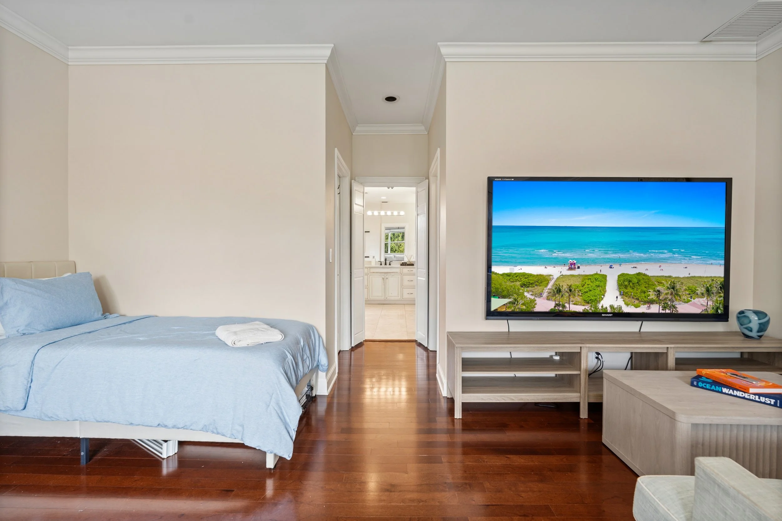 Living room with a bed on the left, a large flat-screen TV displaying a beach scene, and a wooden floor.