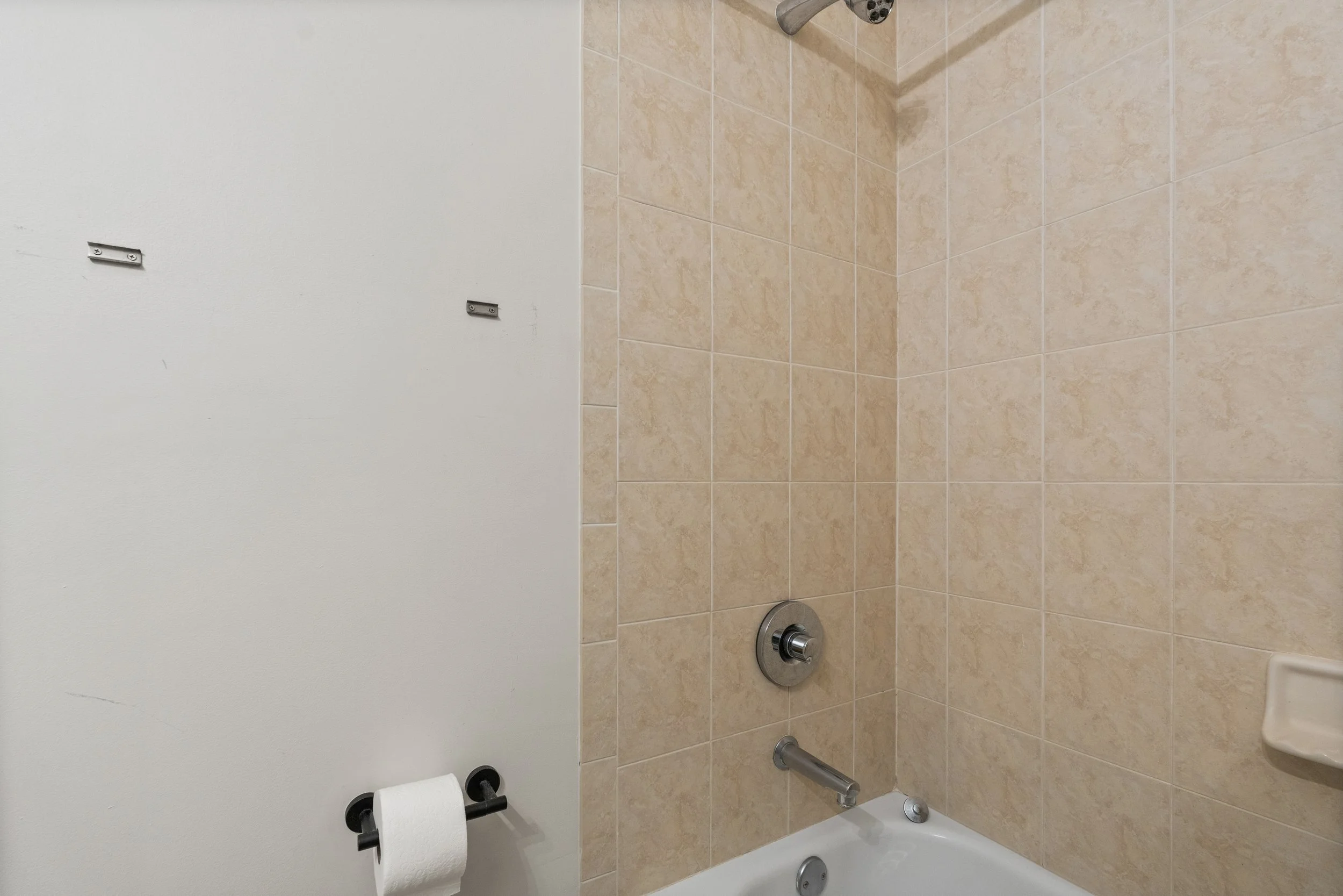 A shower area with beige tiled walls, a chrome showerhead, a bathtub with a rainfall showerhead, and a black toilet paper holder with a roll of toilet paper.