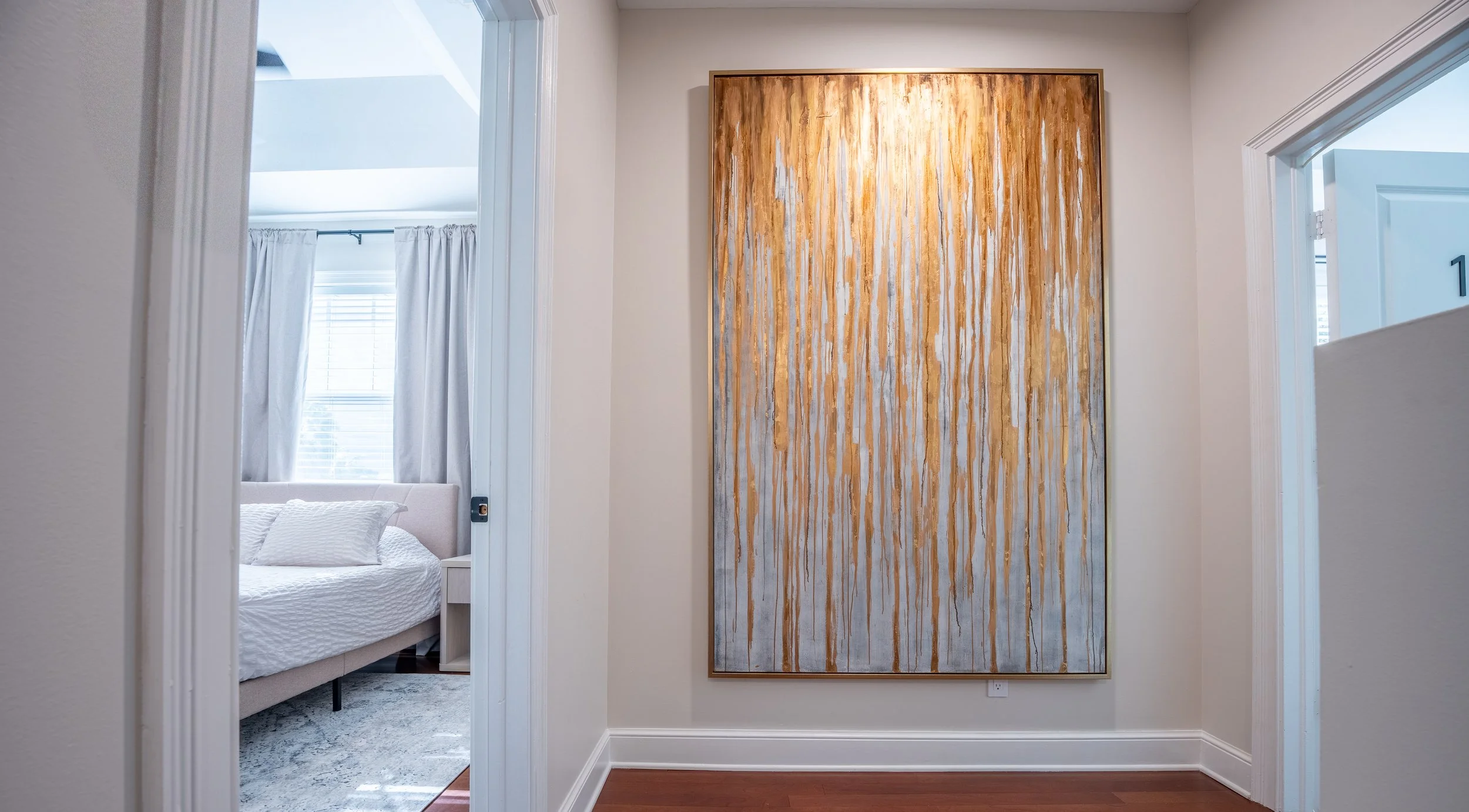 A hallway with a large vertical abstract painting with orange and white streaks on the wall. To the left, a bedroom with a bed, white bedding, and a window with white curtains is visible through an open doorway. To the right, part of a door with the 