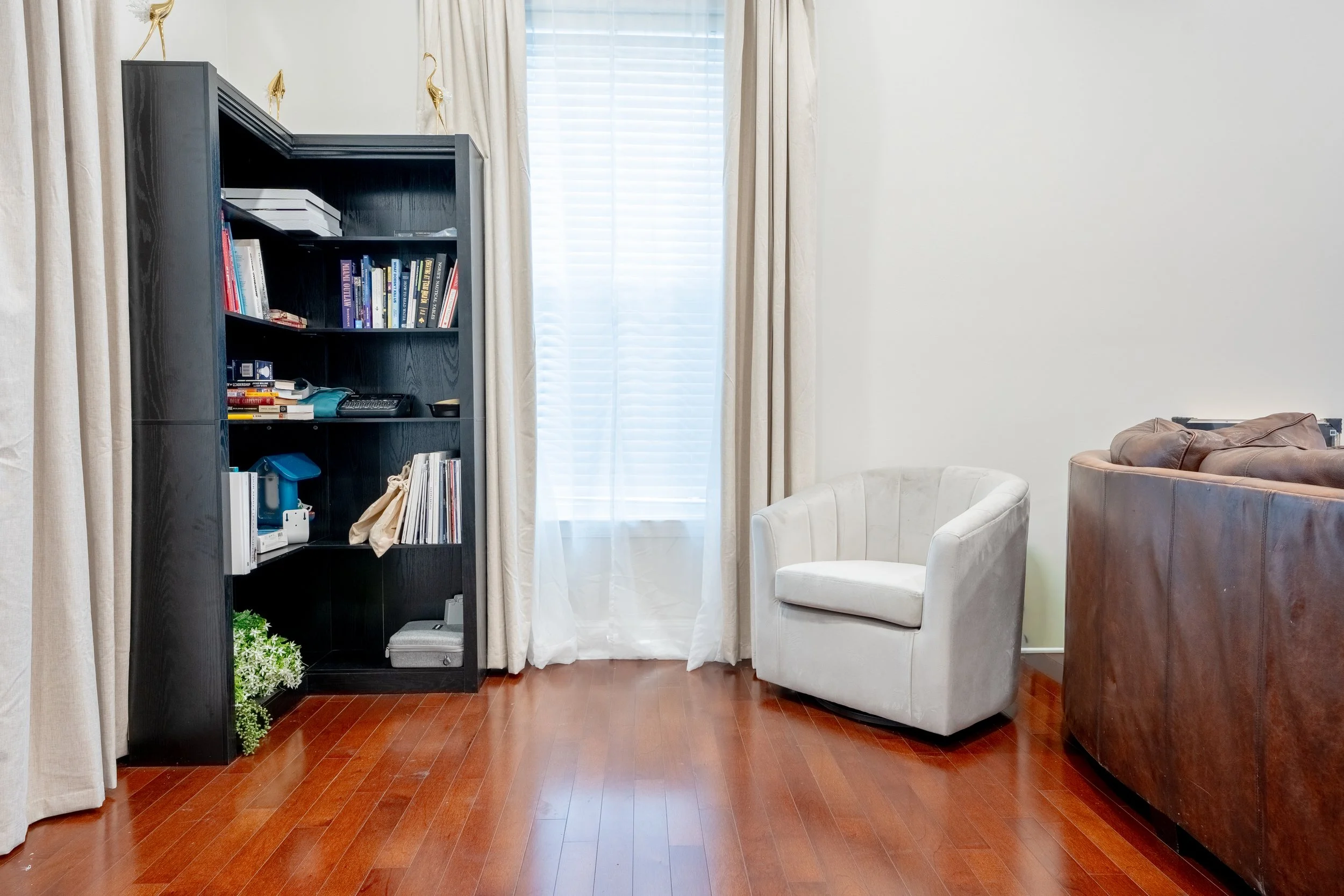 Living room with white walls, a large window with blinds and beige curtains, a black bookshelf filled with books and items, two beige and brown chairs, and hardwood floors.
