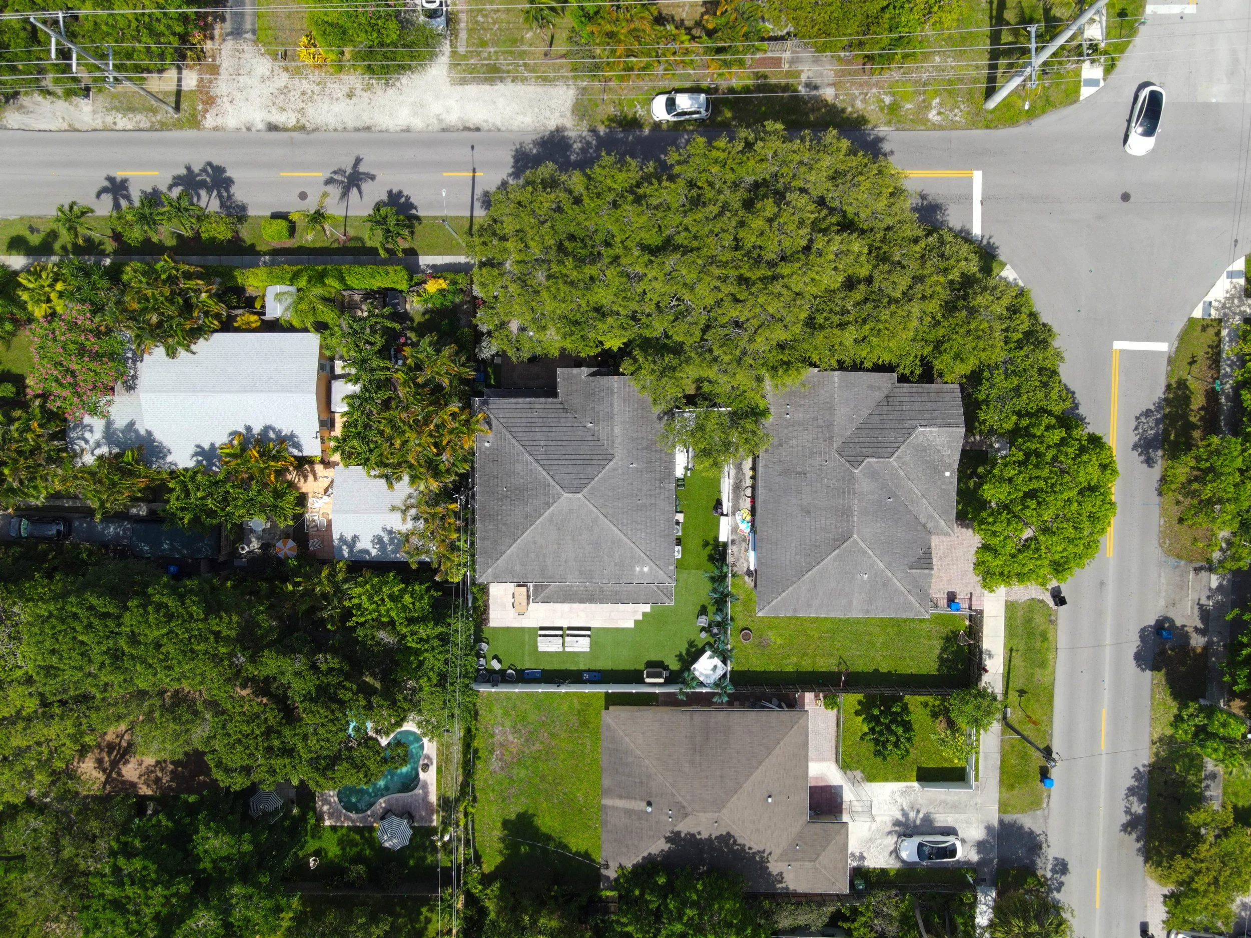 An aerial view of a residential street with houses, trees, automobiles parked along the road, and a swimming pool in someone’s backyard.