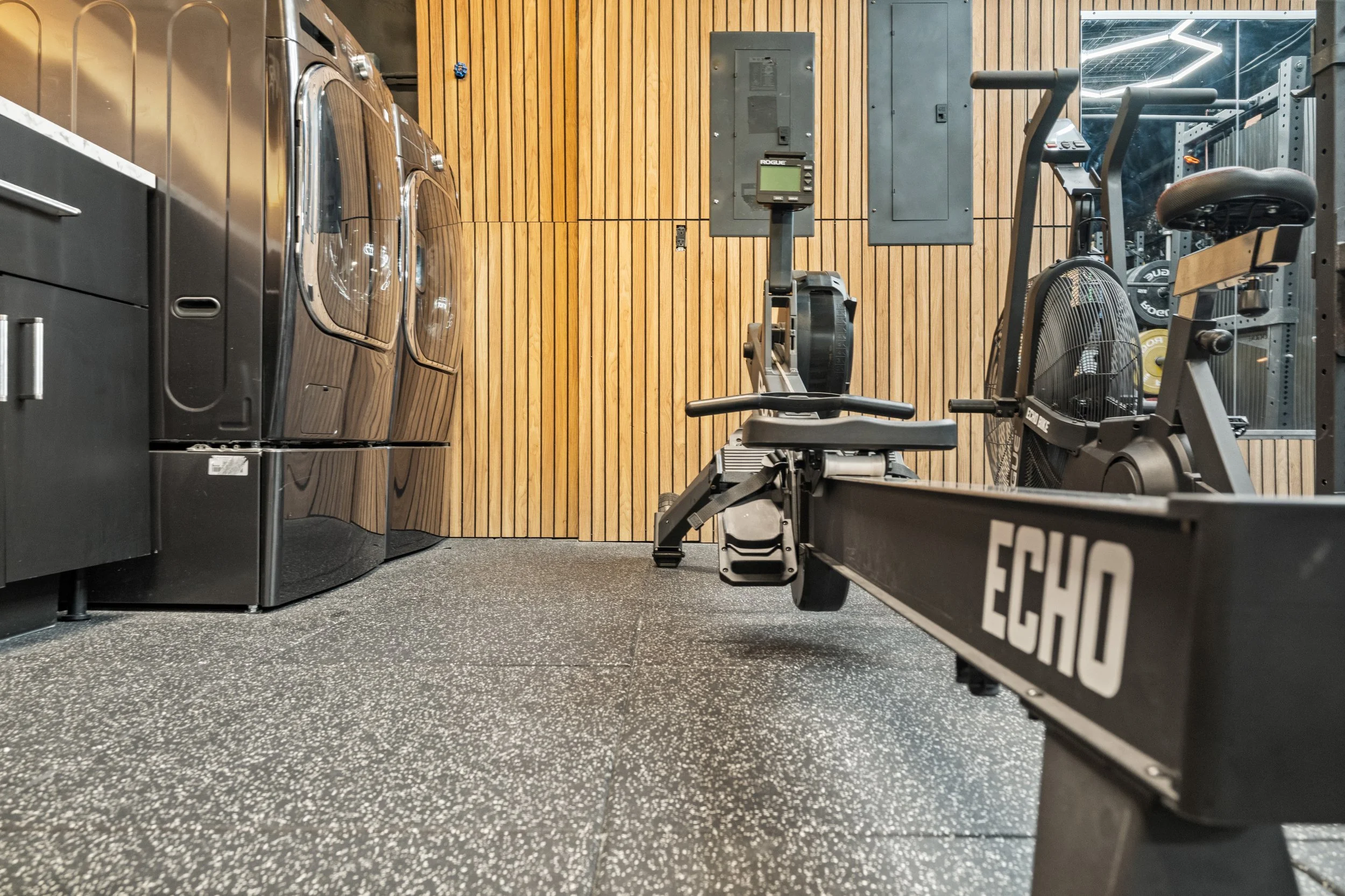 Home gym with wood-paneled wall, washer and dryer on the left, with exercise equipment including an elliptical trainer and a bike in the center and right.