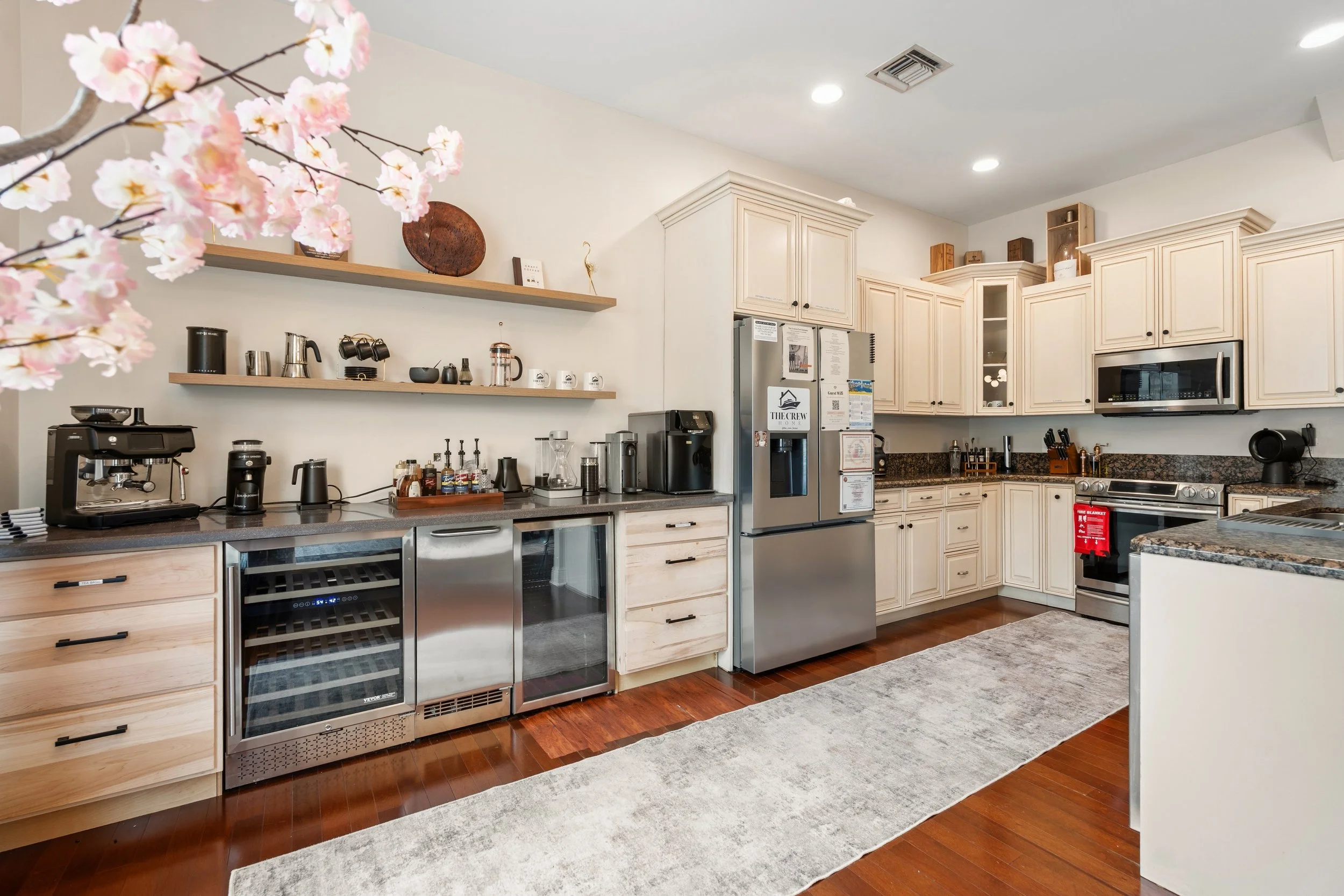 A kitchen with cream-colored cabinets, granite countertops, stainless steel appliances, open wooden shelves with decorative items, and pink cherry blossom flowers in the foreground.