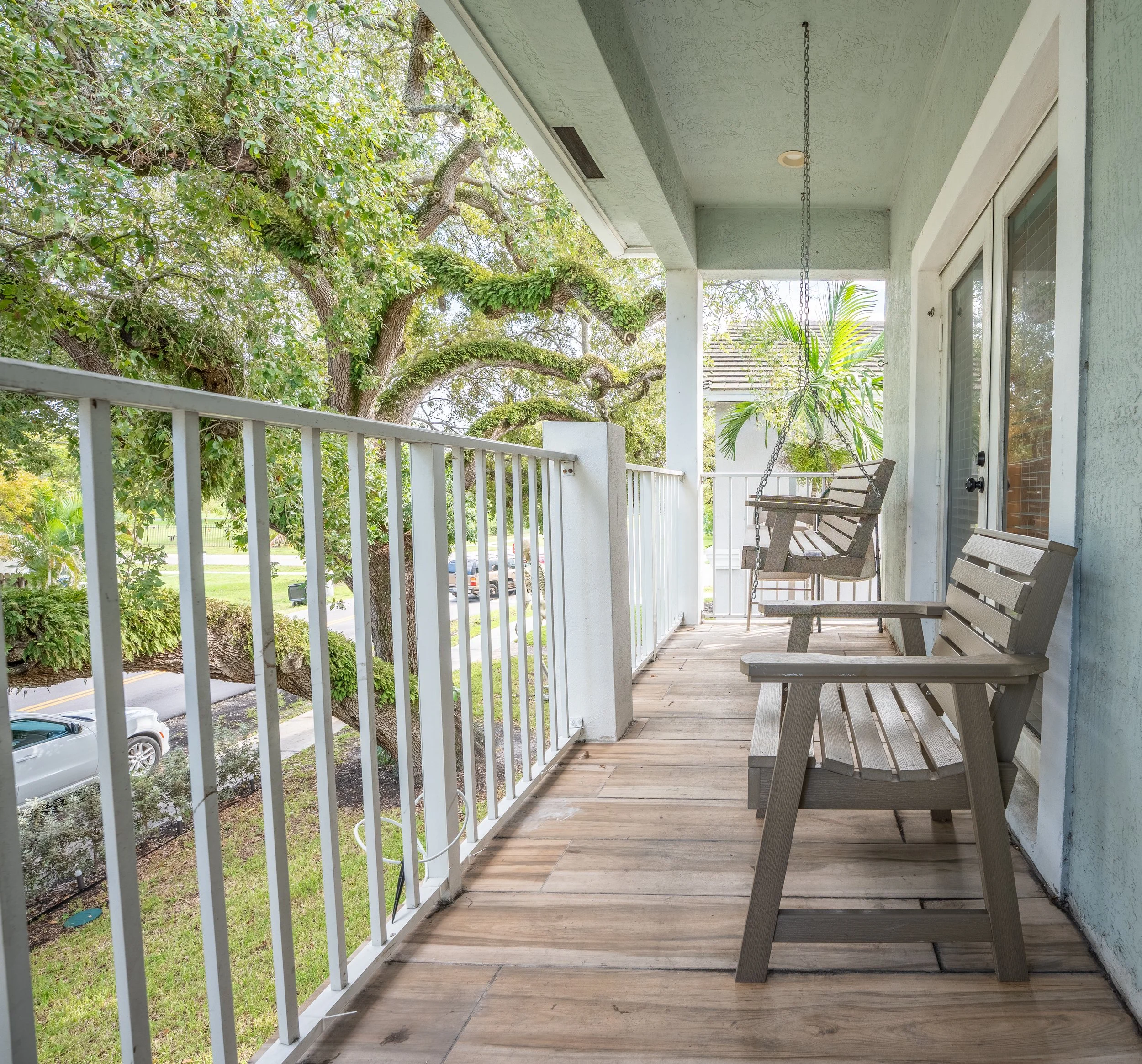 A porch with a white railing and wooden floor, two wooden benches, an outdoor swing hanging from the ceiling, and a view of trees and a street with cars.
