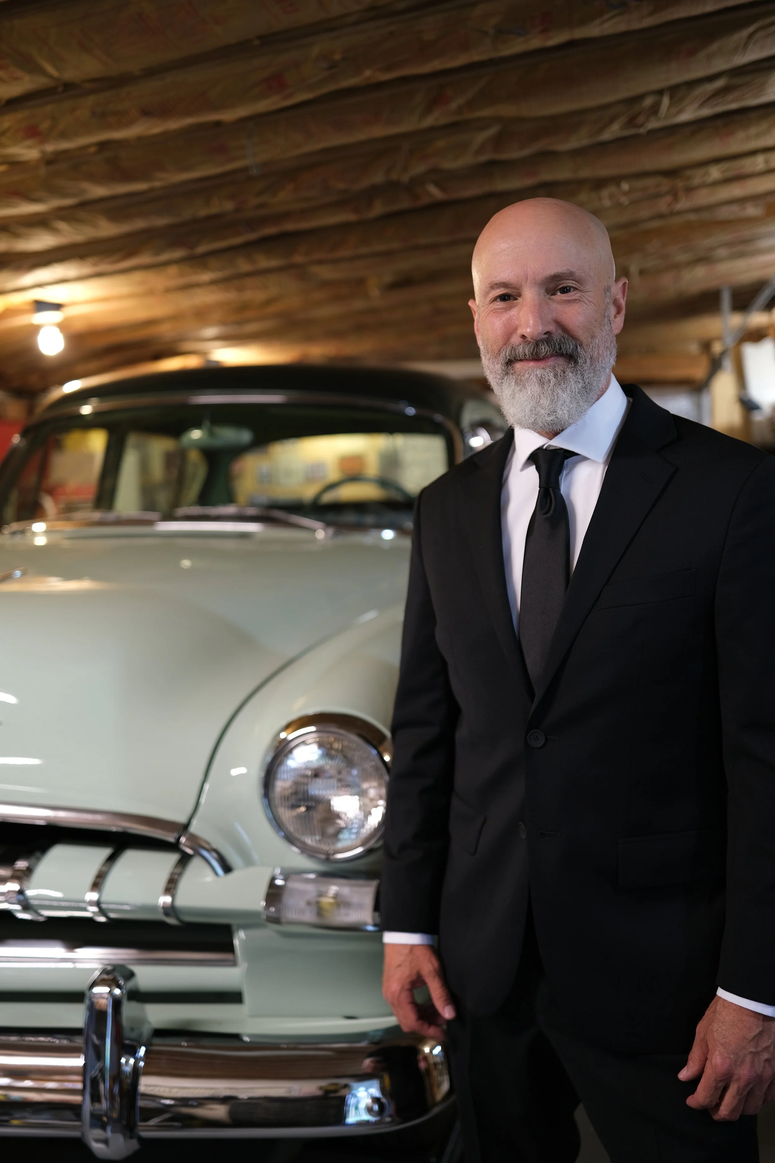 A friendly and handsome man in a sharp black suit and tie standing next to a vintage light green car, a fully restored, 1953 Plymouth Cranbrook inside a garage with a wooden ceiling.