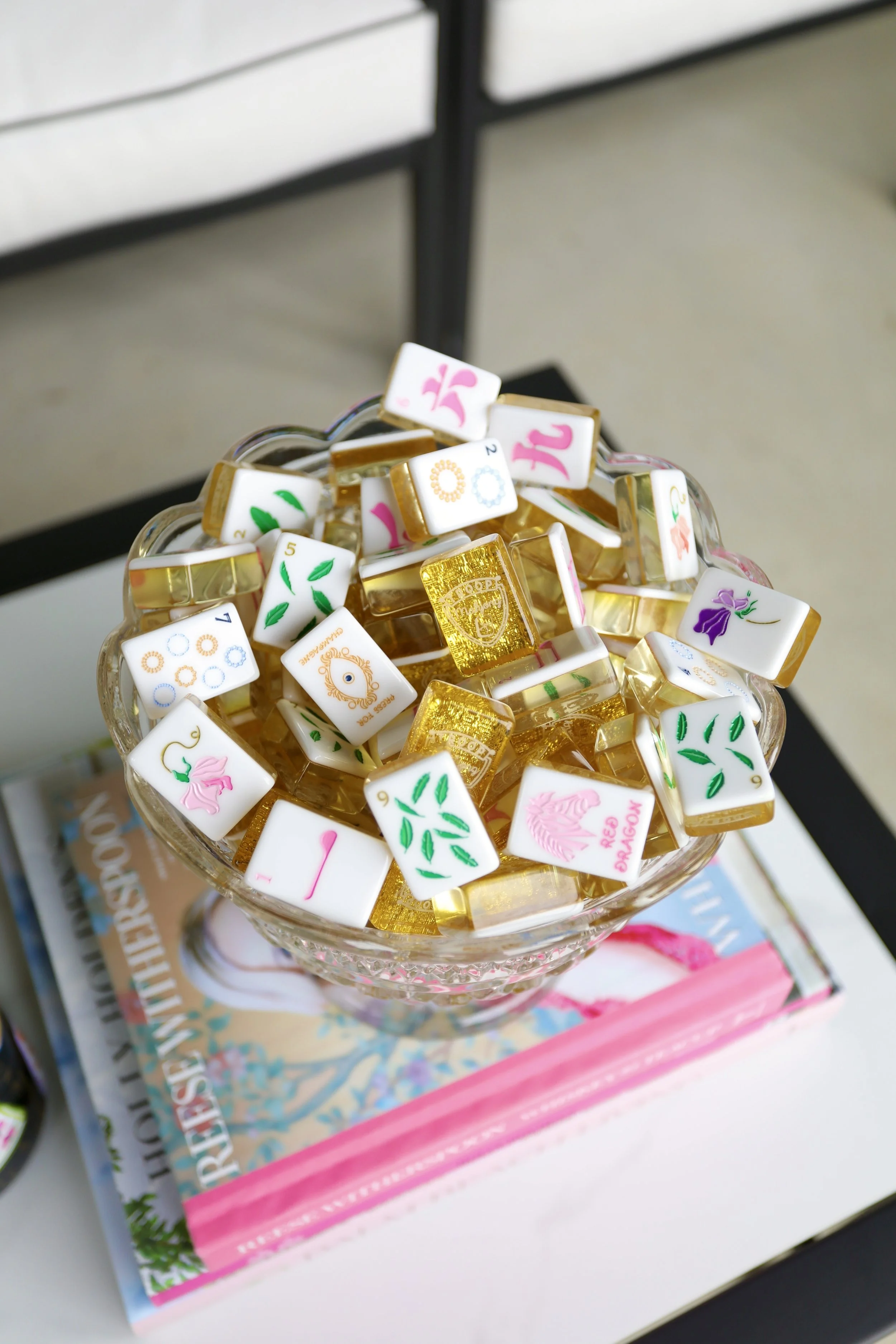 A glass bowl filled with decorative domino tiles, including gold, white, and colorful tiles with floral and circular designs, placed on top of books and magazines on a white surface.