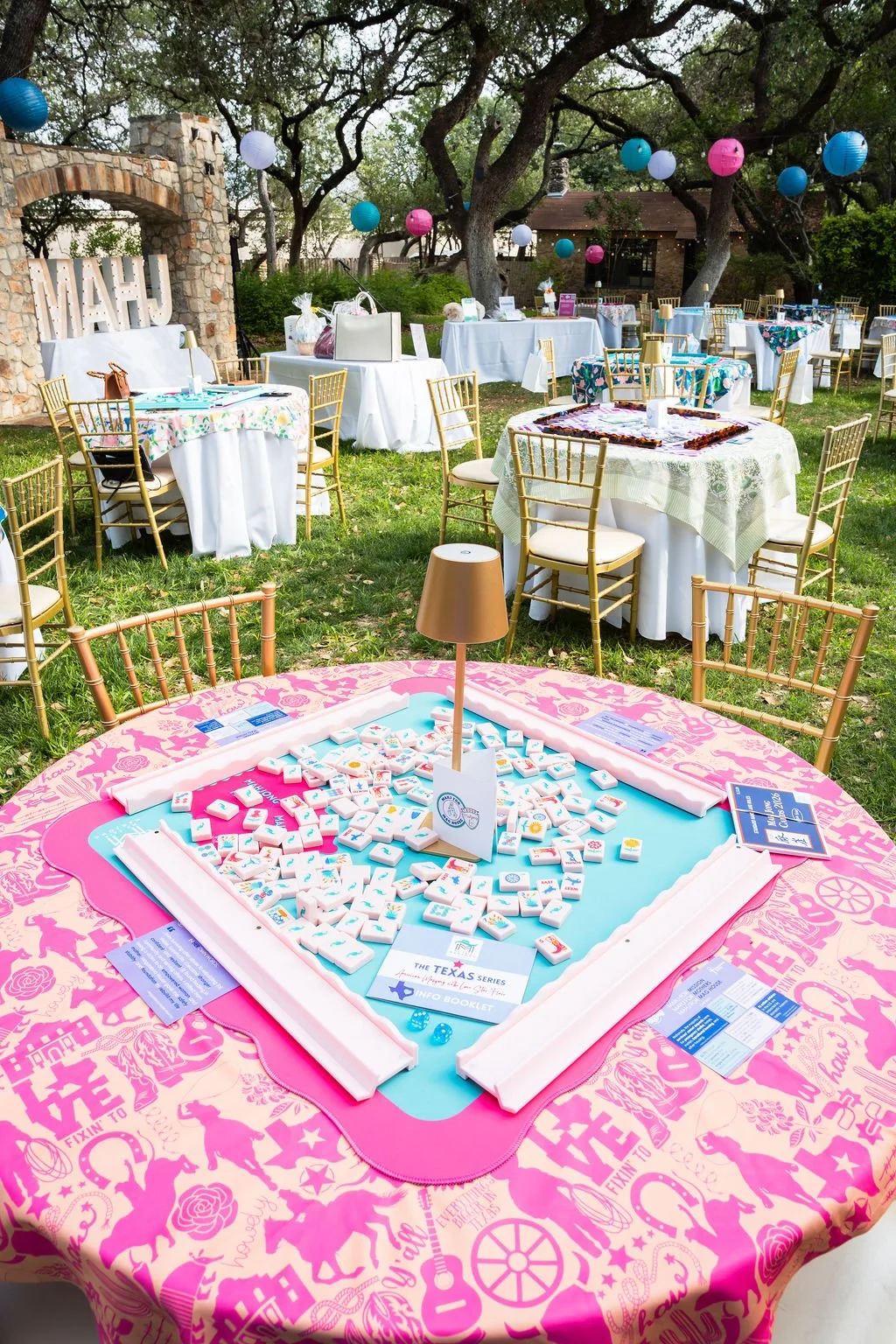 Outdoor bridal shower or celebration with tables decorated with colorful tablecloths, gold chairs, and balloon decorations hanging from trees, featuring a domino game setup on a pink tablecloth in the foreground.